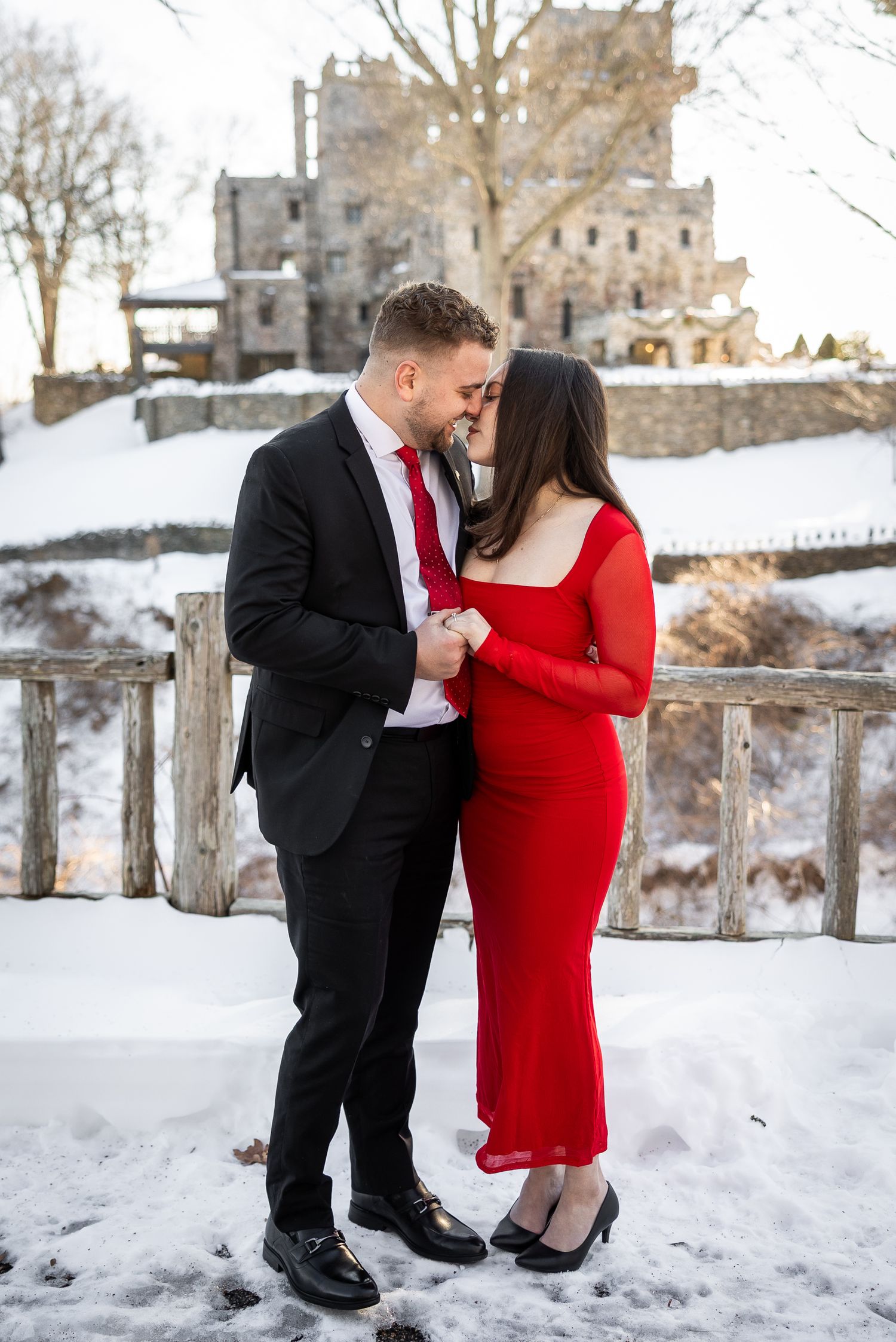 A couple in formal attire poses together in a snowy winter setting, with the person in red kneeling down.