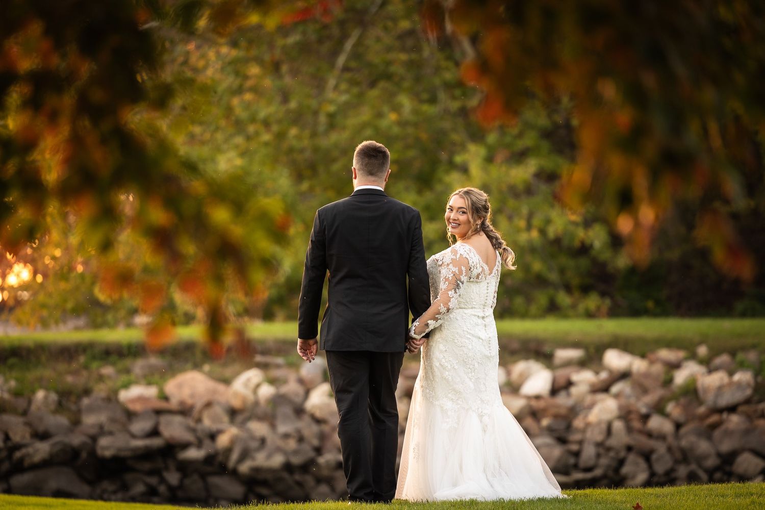 A couple walks hand in hand through a beautiful autumn landscape with golden leaves and stone walls.
