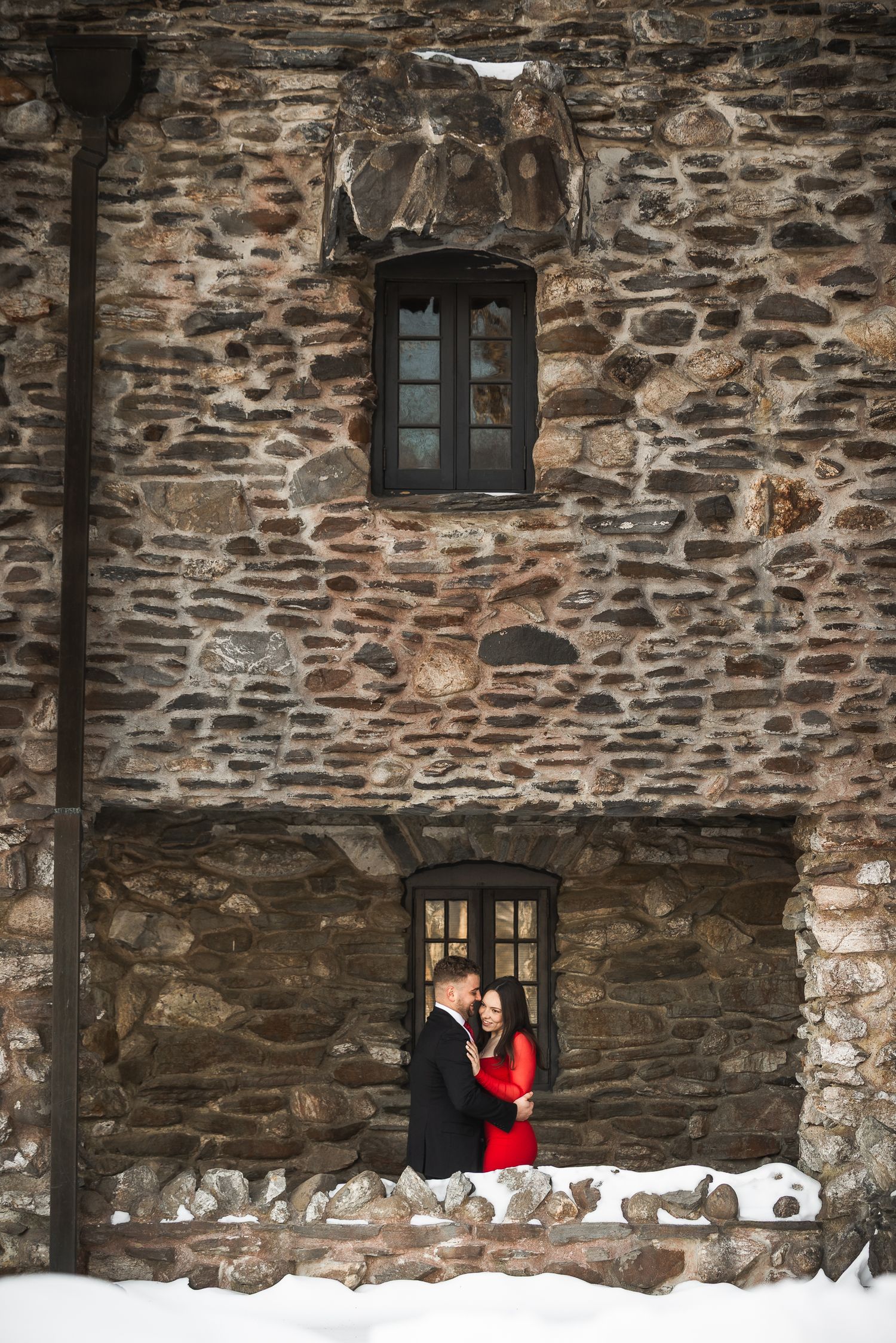 Two people stand beside a historic stone building with dark windows during a snowy winter day.
