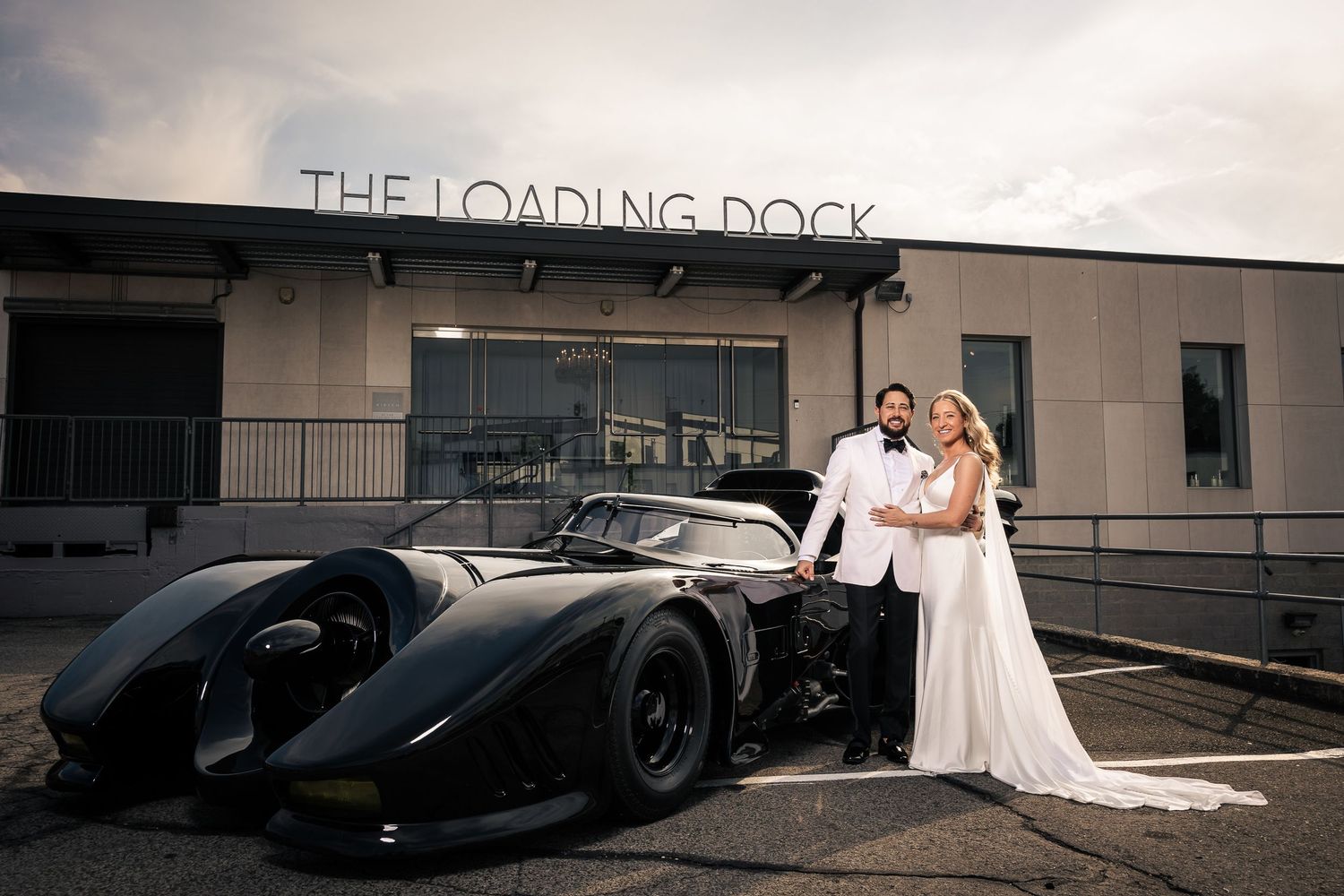 A wedding couple poses with a sleek black vintage-style sports car outside The Loading Dock building.