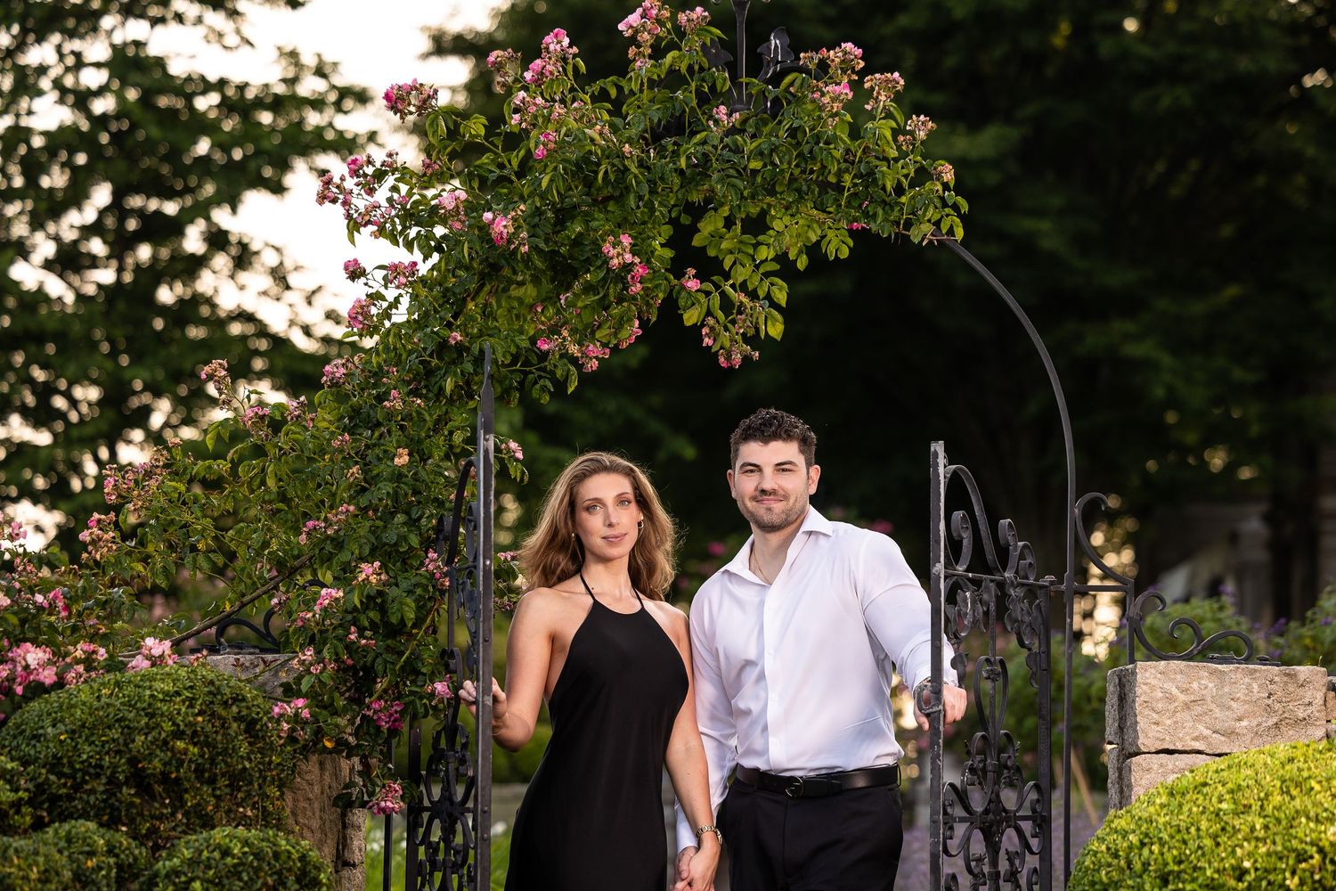 A couple in formal attire shares a romantic moment in a lush garden setting with blooming flowers and greenery.