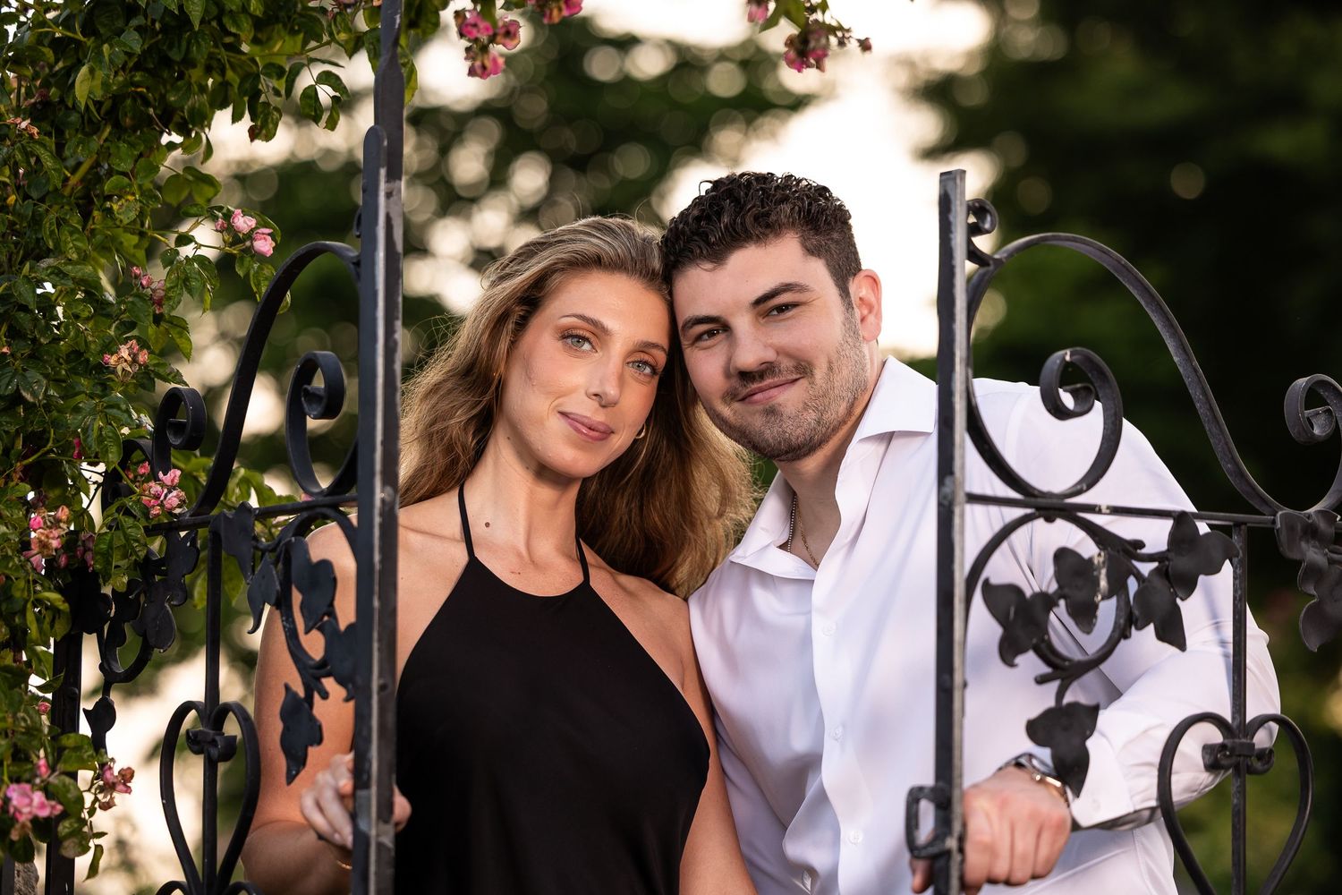 A couple in black and white attire pose together outdoors near decorative iron railings and greenery.