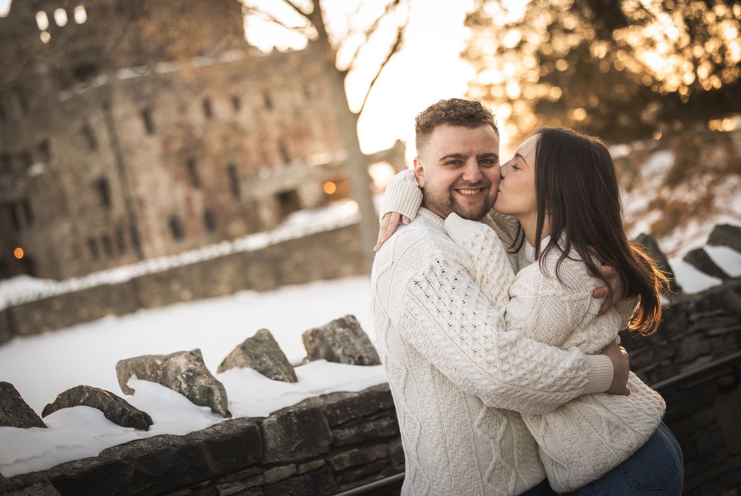 A couple in cozy white sweaters embraces outdoors during a snowy winter sunset.