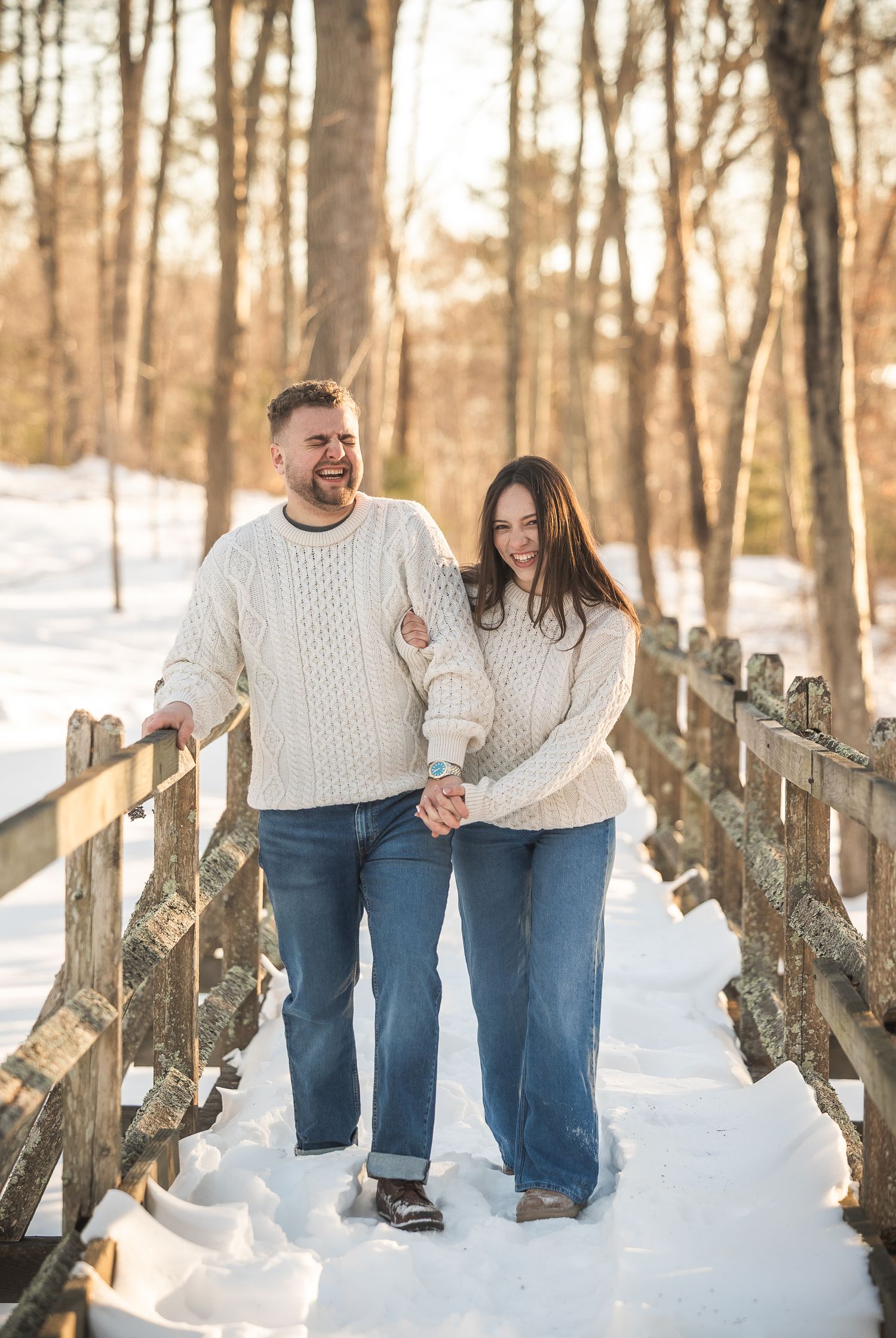 A couple in matching white sweaters walks together on a snowy path between wooden fences in a winter forest.