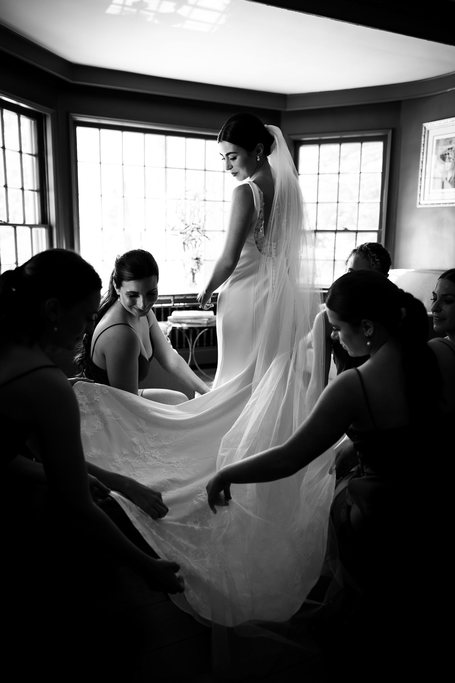 Wedding dress being adjusted by helpers in dramatic black and white lighting.