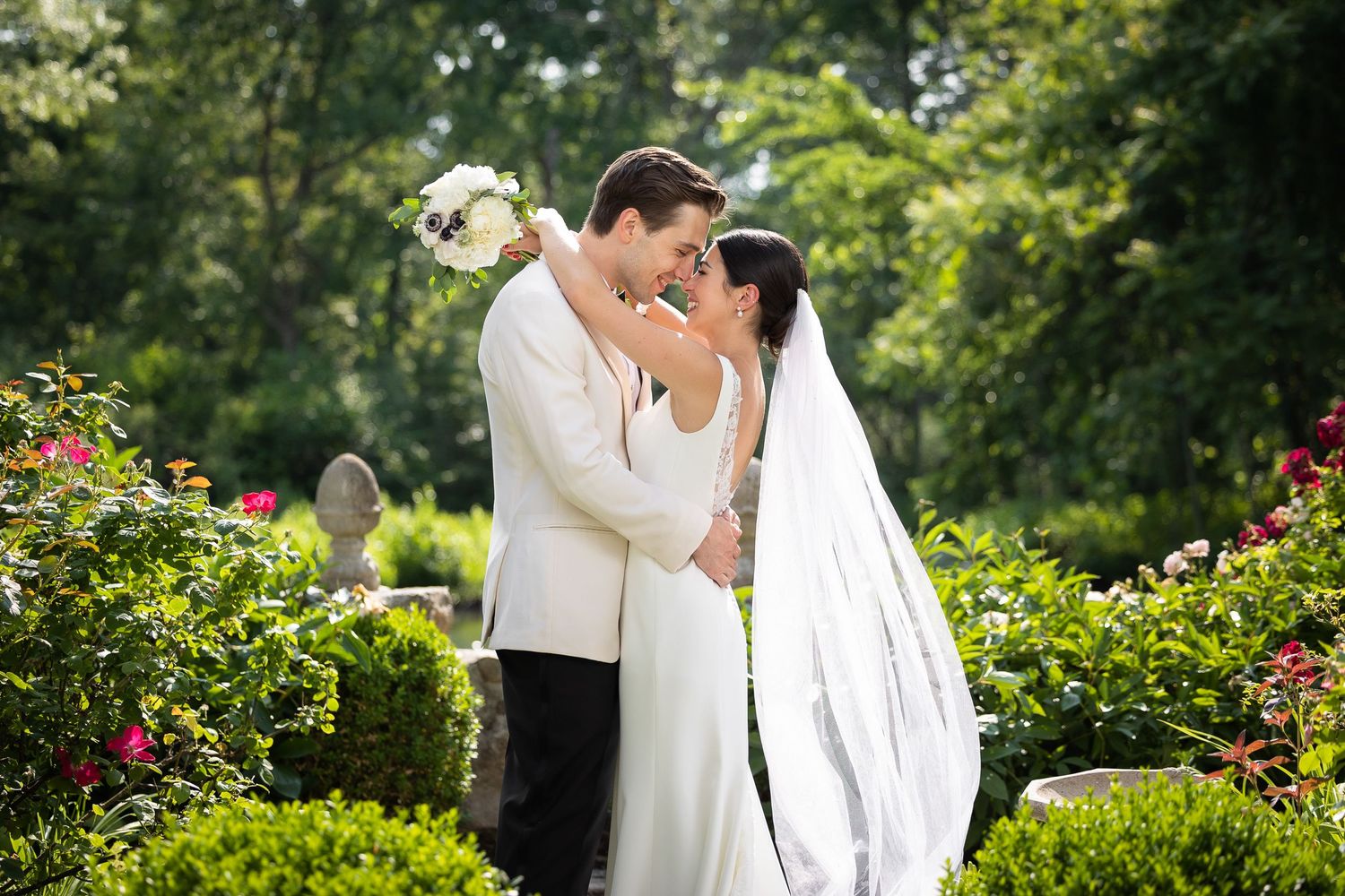 Newlyweds share a romantic kiss in a garden setting surrounded by flowers and greenery.