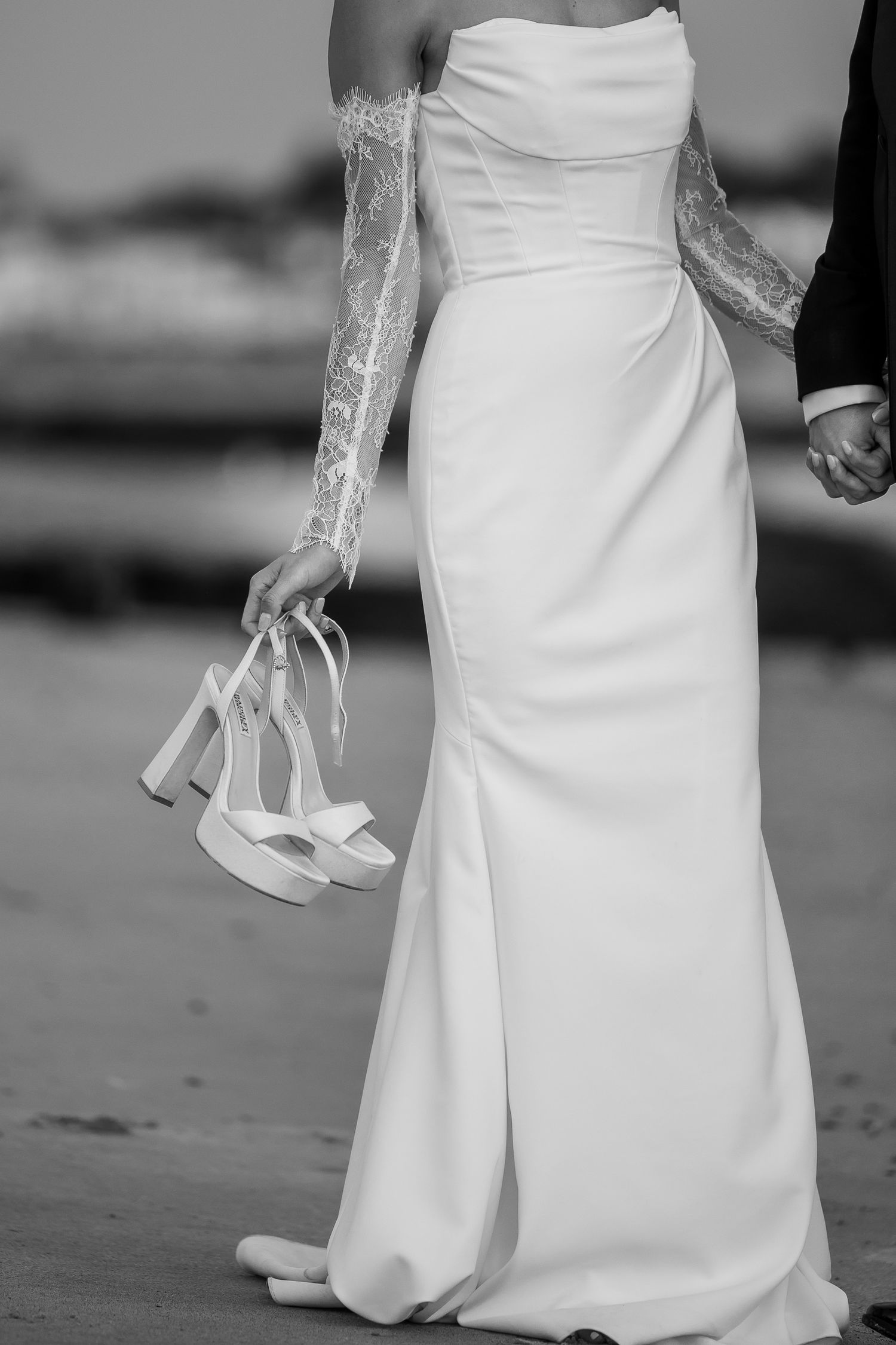 A close-up view of a white wedding dress hem and delicate lace shoes being held in hand against a dark background.
