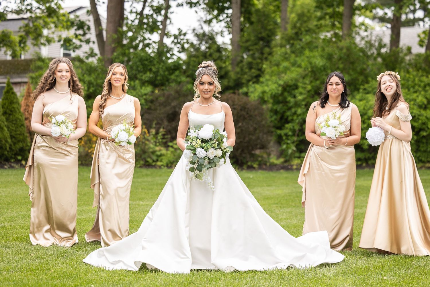Bridal party in champagne dresses poses together on green lawn for formal wedding photos.