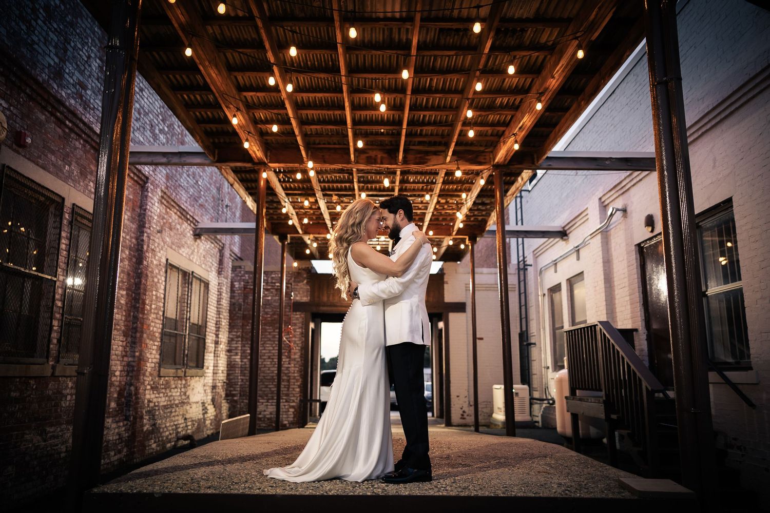 Romantic couple in elegant wedding attire share intimate moments in an industrial urban setting with string lights overhead.