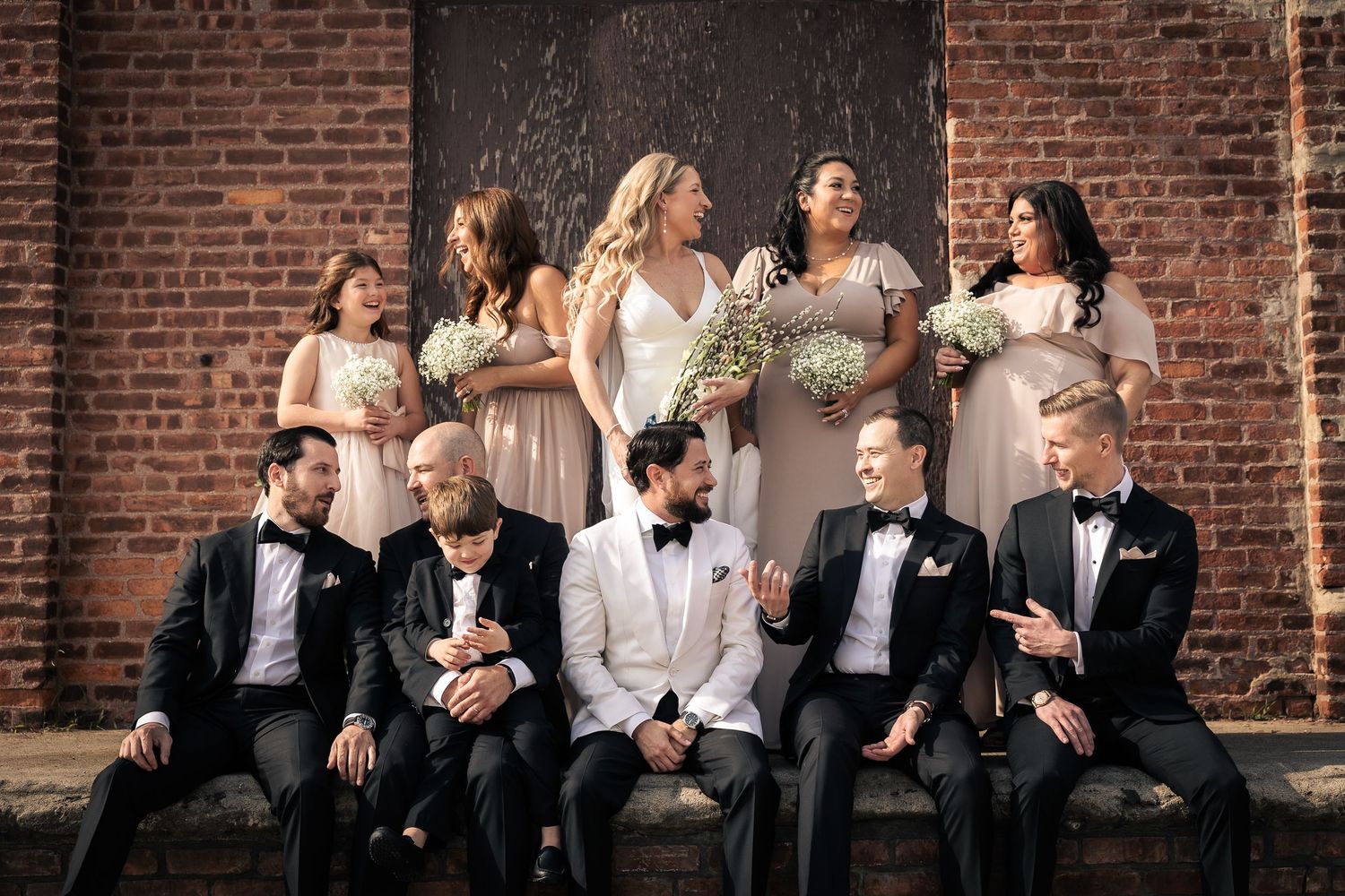 Wedding party in formal attire poses together against rustic brick wall at elegant venue.