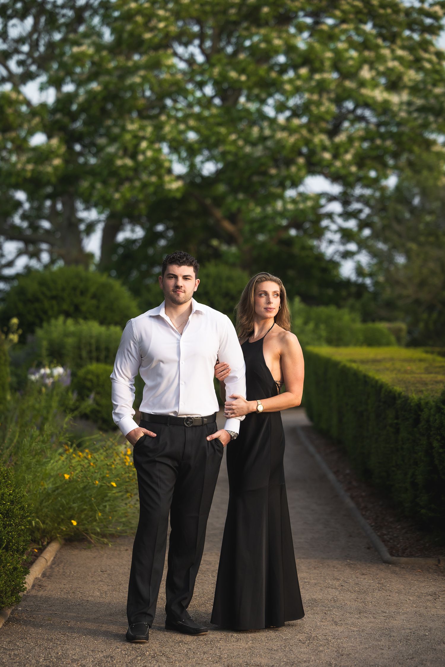 A couple in formal black and white attire poses together on a garden path surrounded by green hedges and trees.