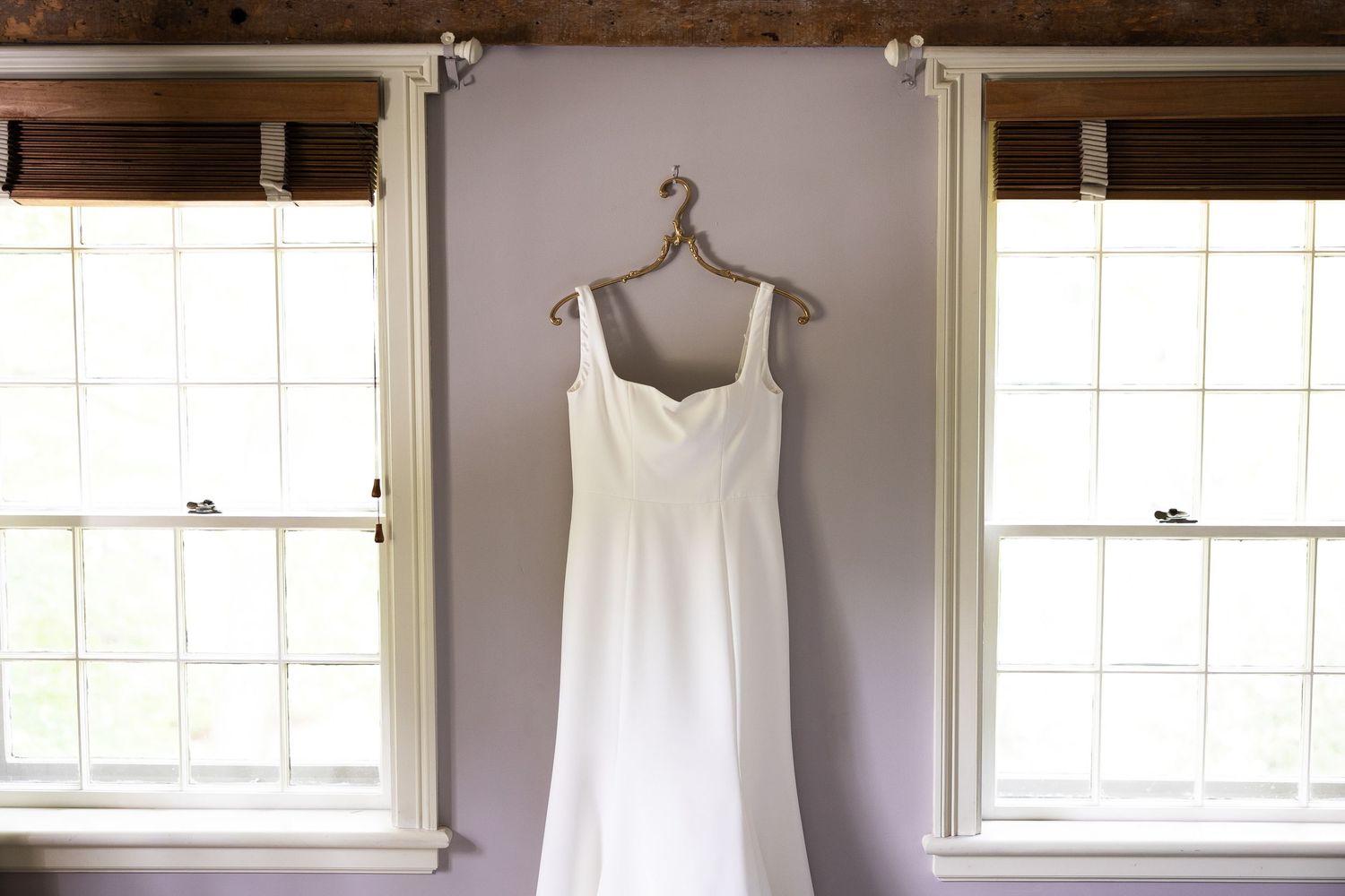 White wedding dress hanging on wooden hangers against a bright window with natural light.