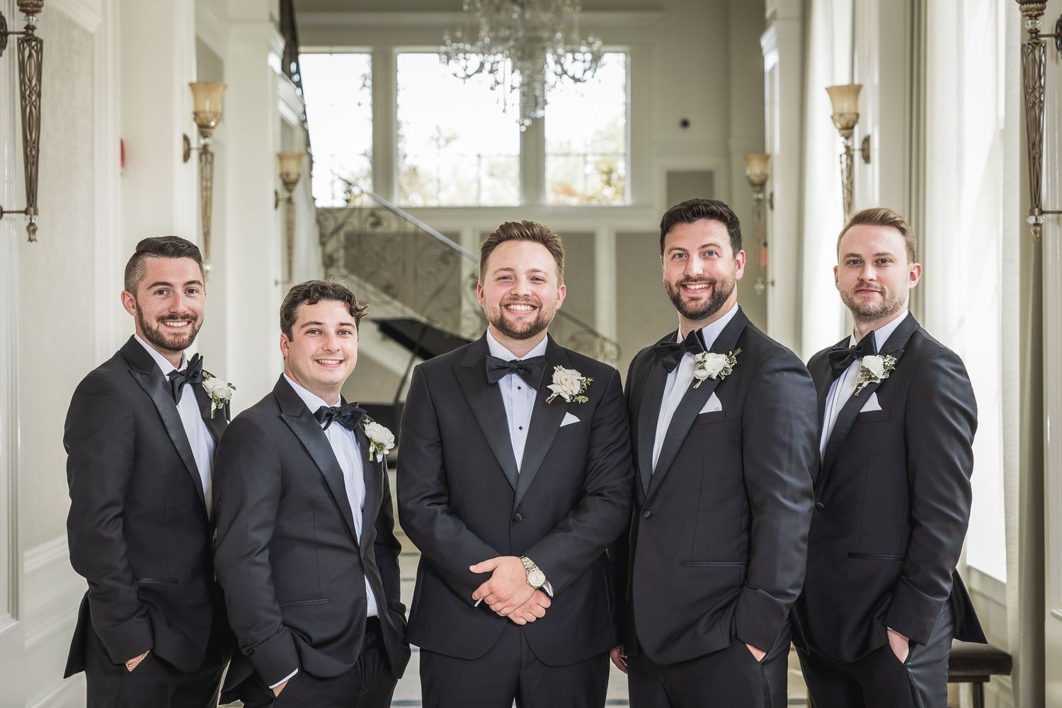 Groomsmen in black tuxedos posing in an elegant white hallway.