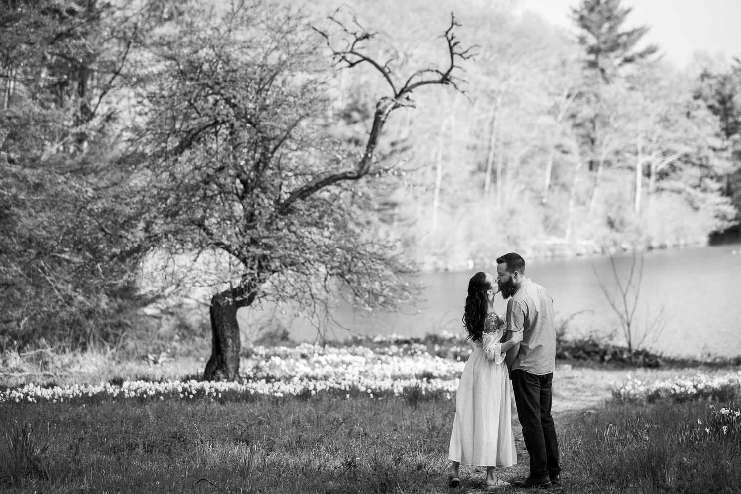 A romantic black and white photo of a couple standing by a lake under a twisted tree in springtime.