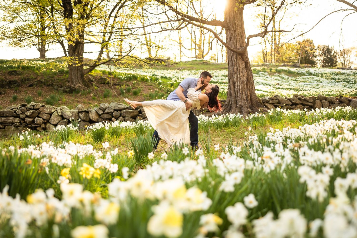 Two people sit together among blooming white daffodils in a spring garden during golden hour sunset.