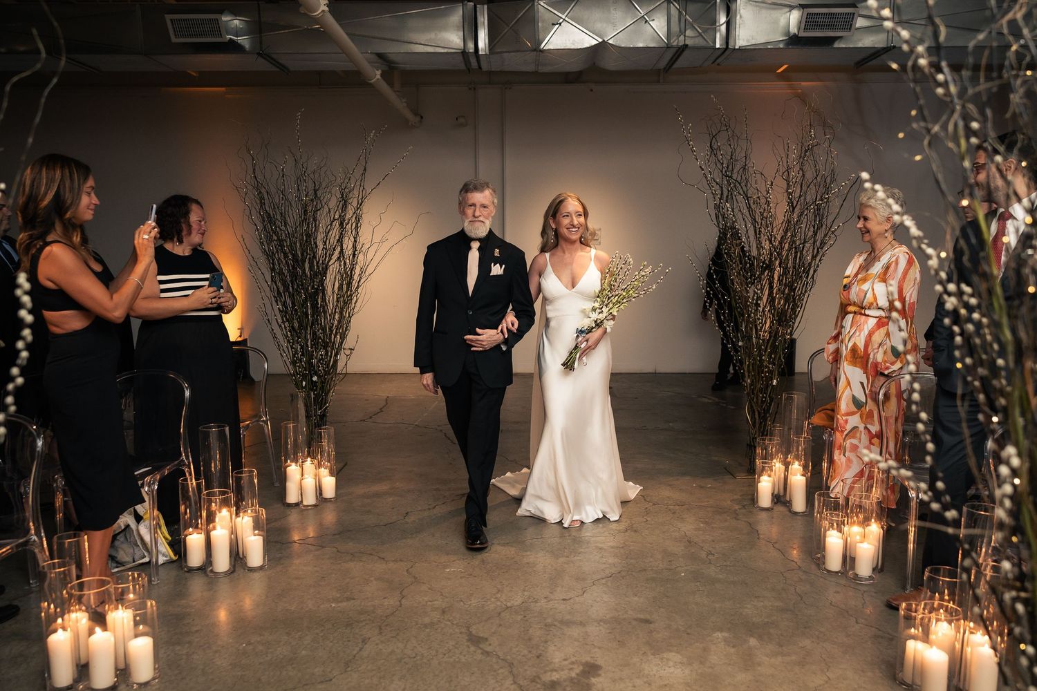 A couple walks down a candlelit aisle decorated with tall branches and twinkling lights during an indoor ceremony.