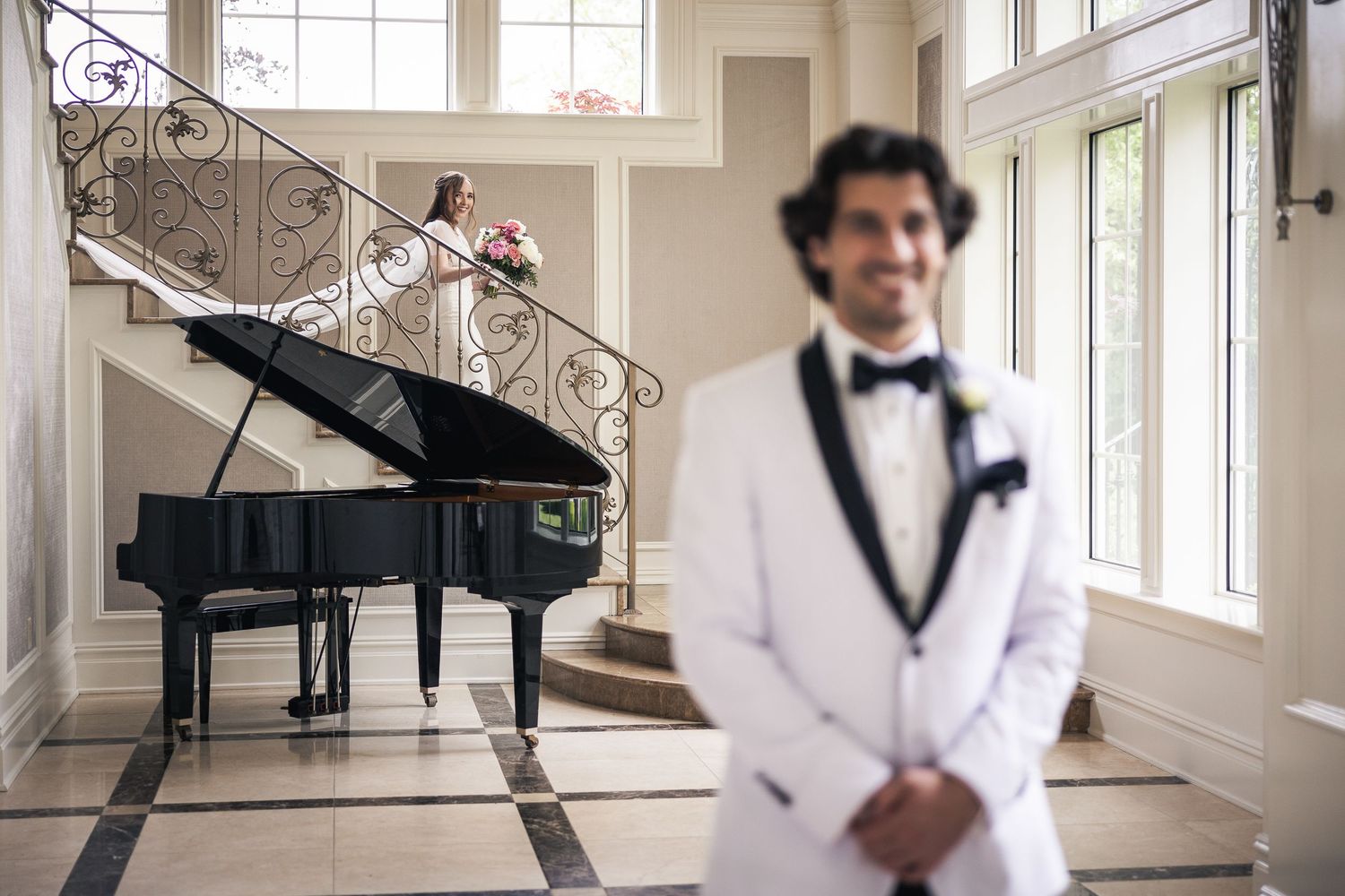 A person in a black tuxedo poses elegantly in a bright white room with large windows.