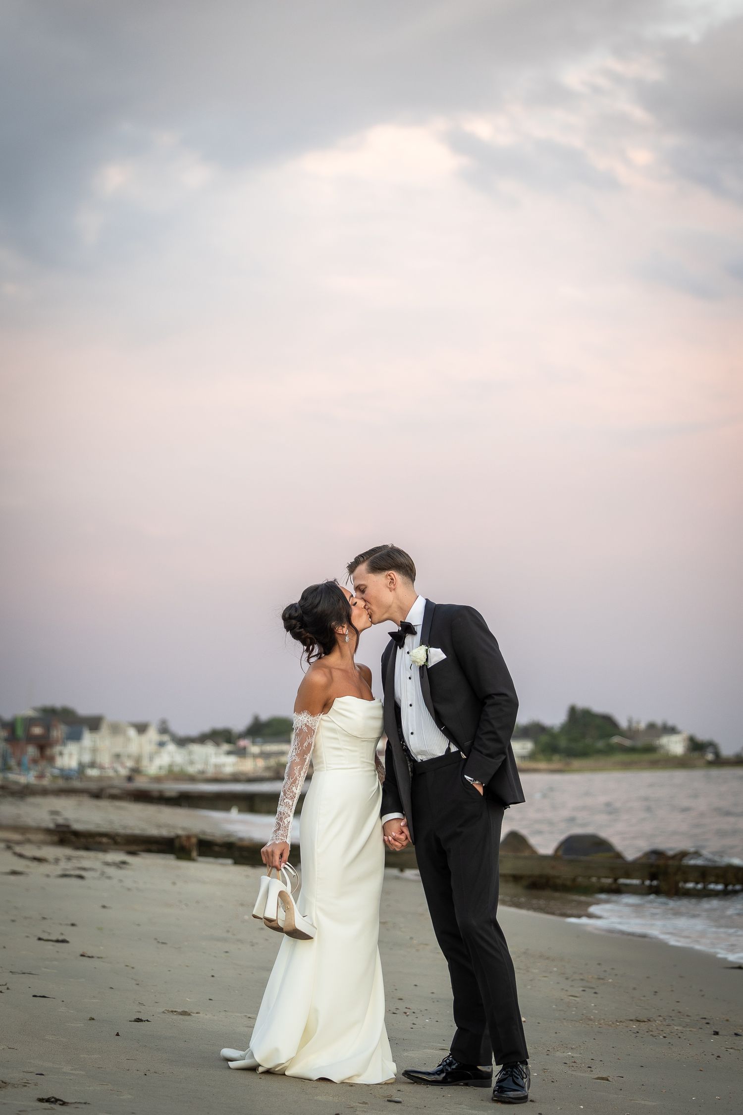Newlyweds share romantic moment on beach at sunset with coastal homes in background.