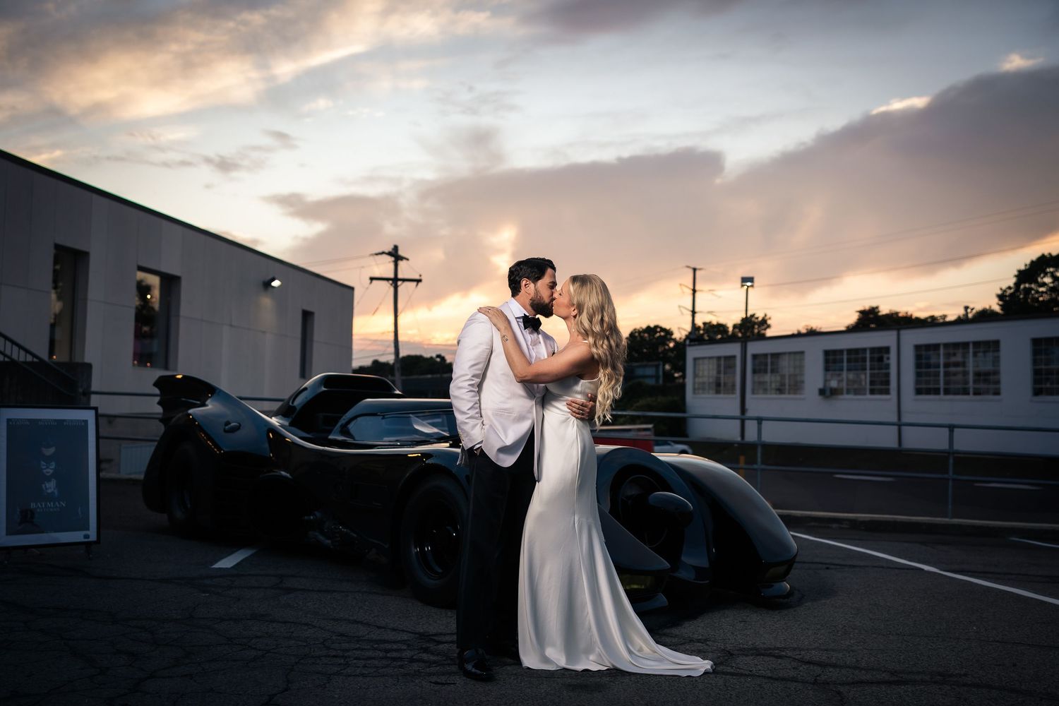 A couple embraces in a parking lot at sunset with a classic car in the background.