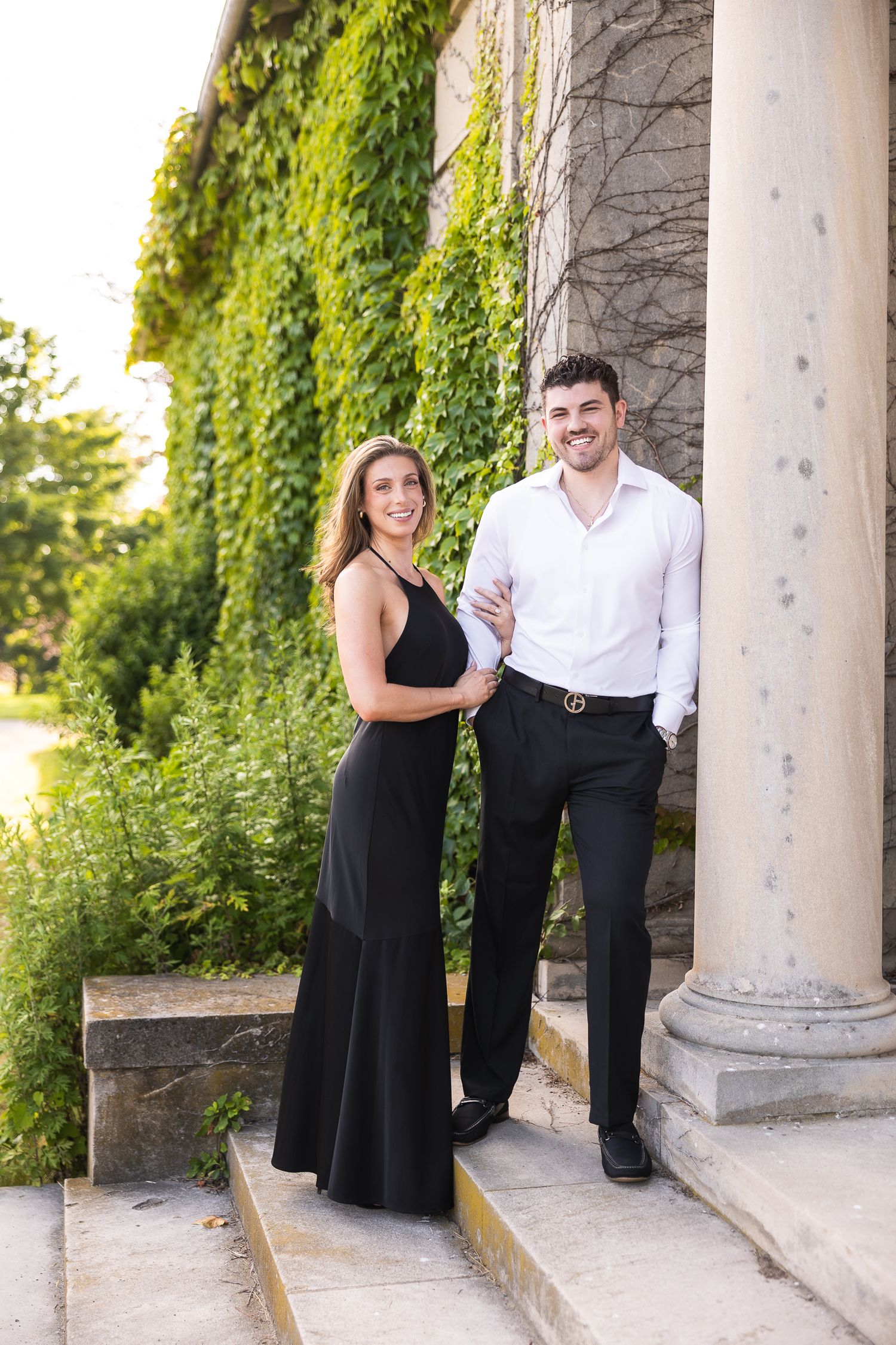 Couple in black and white attire share romantic moments under stone columns with green ivy backdrop.