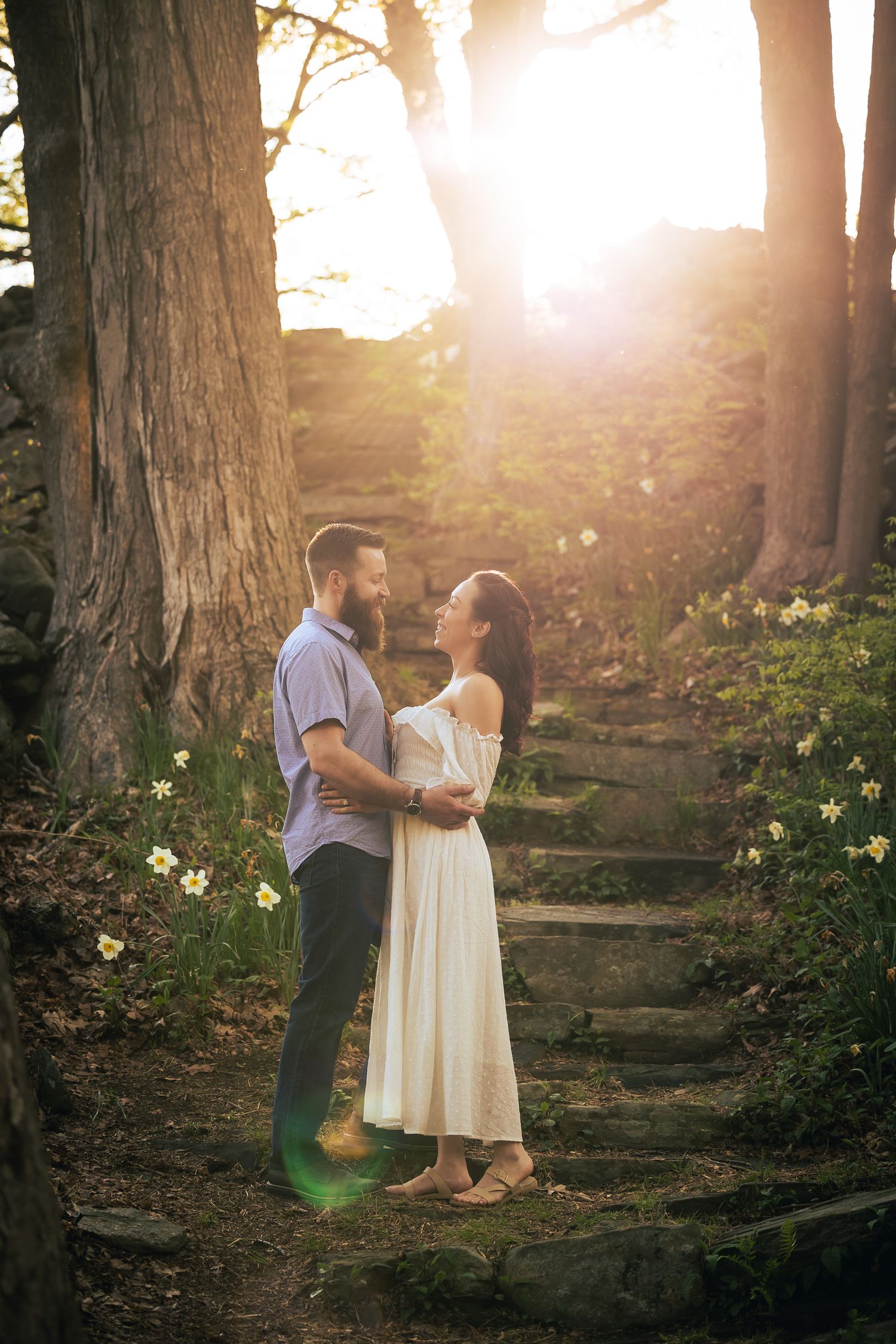 A couple shares a romantic moment on stone steps in a sunlit forest setting during golden hour.