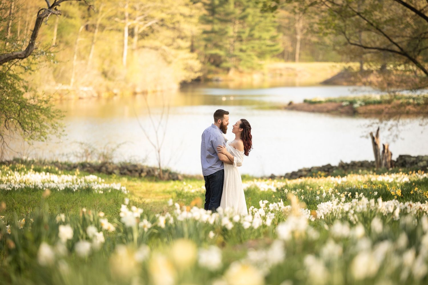 A couple embraces in a spring garden filled with blooming white daffodils beside a tranquil lake at sunset.