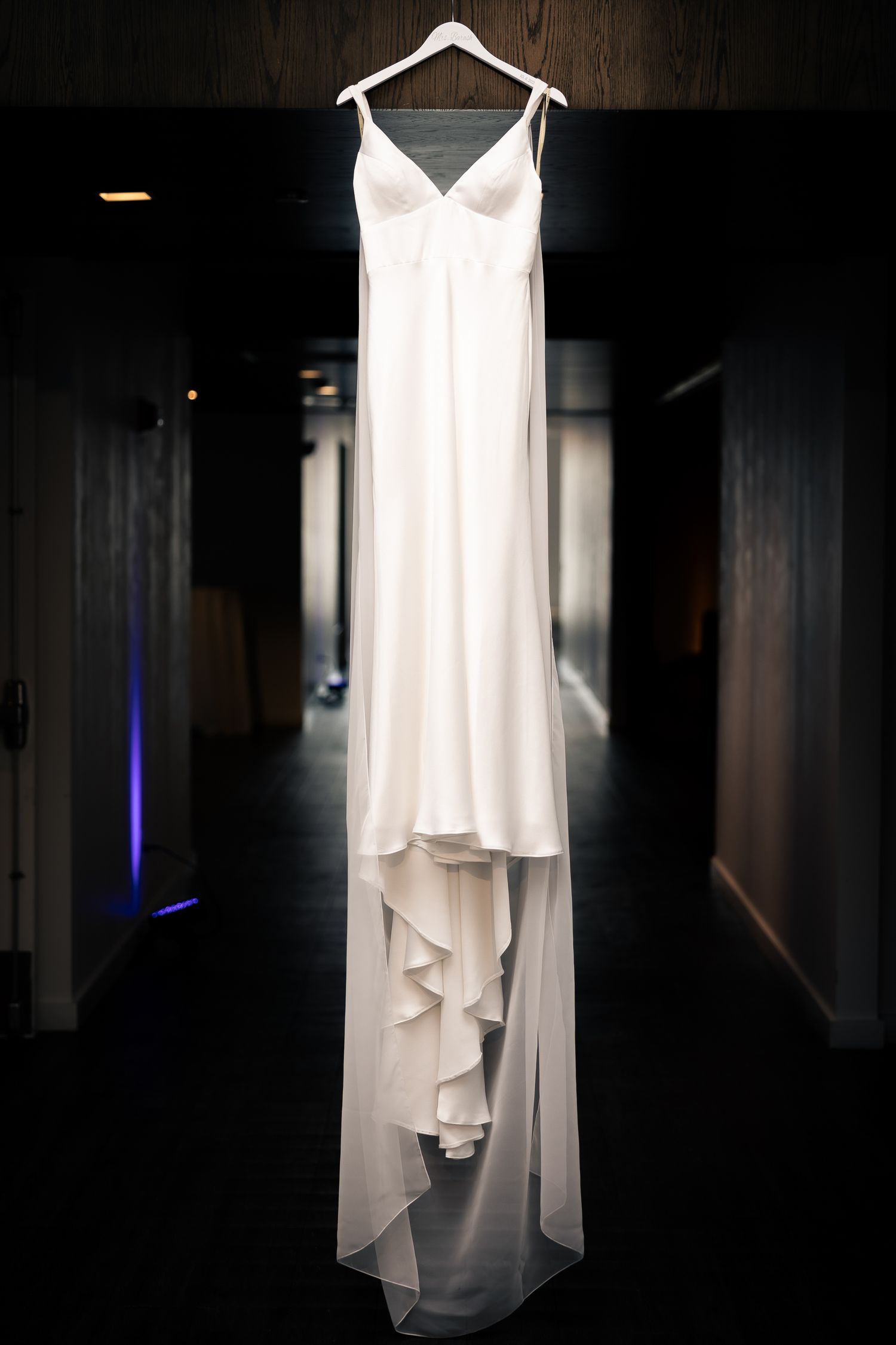 A white wedding dress with flowing layers hangs elegantly against a dark wooden background.