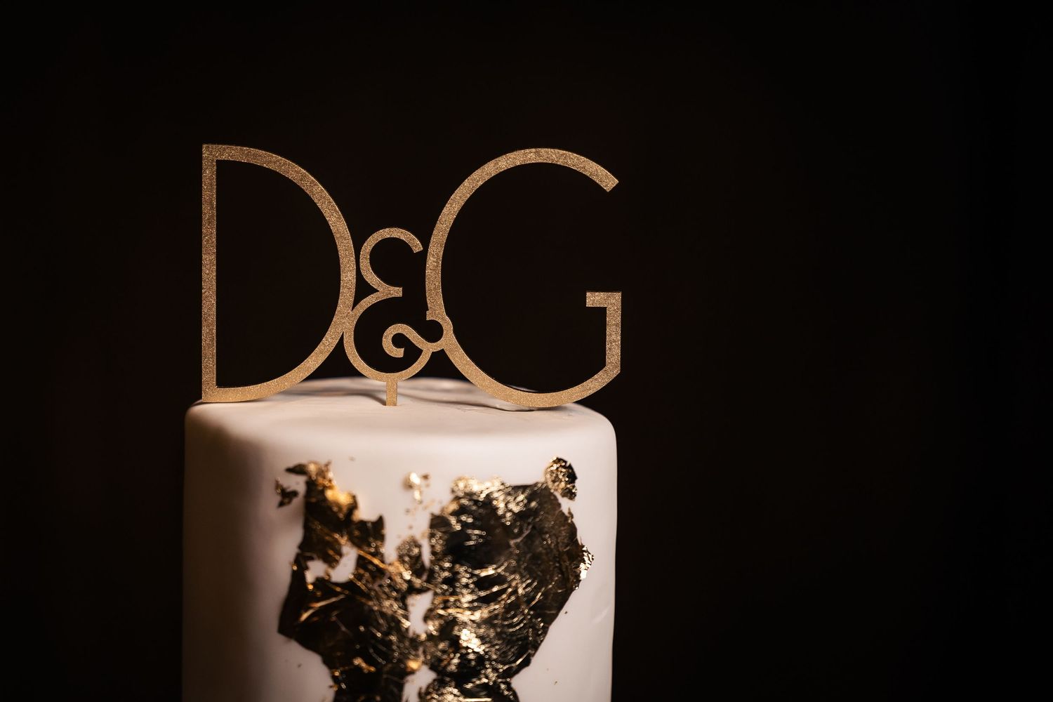 A white wedding cake decorated with elegant gold details and black accents sits against a dark background.