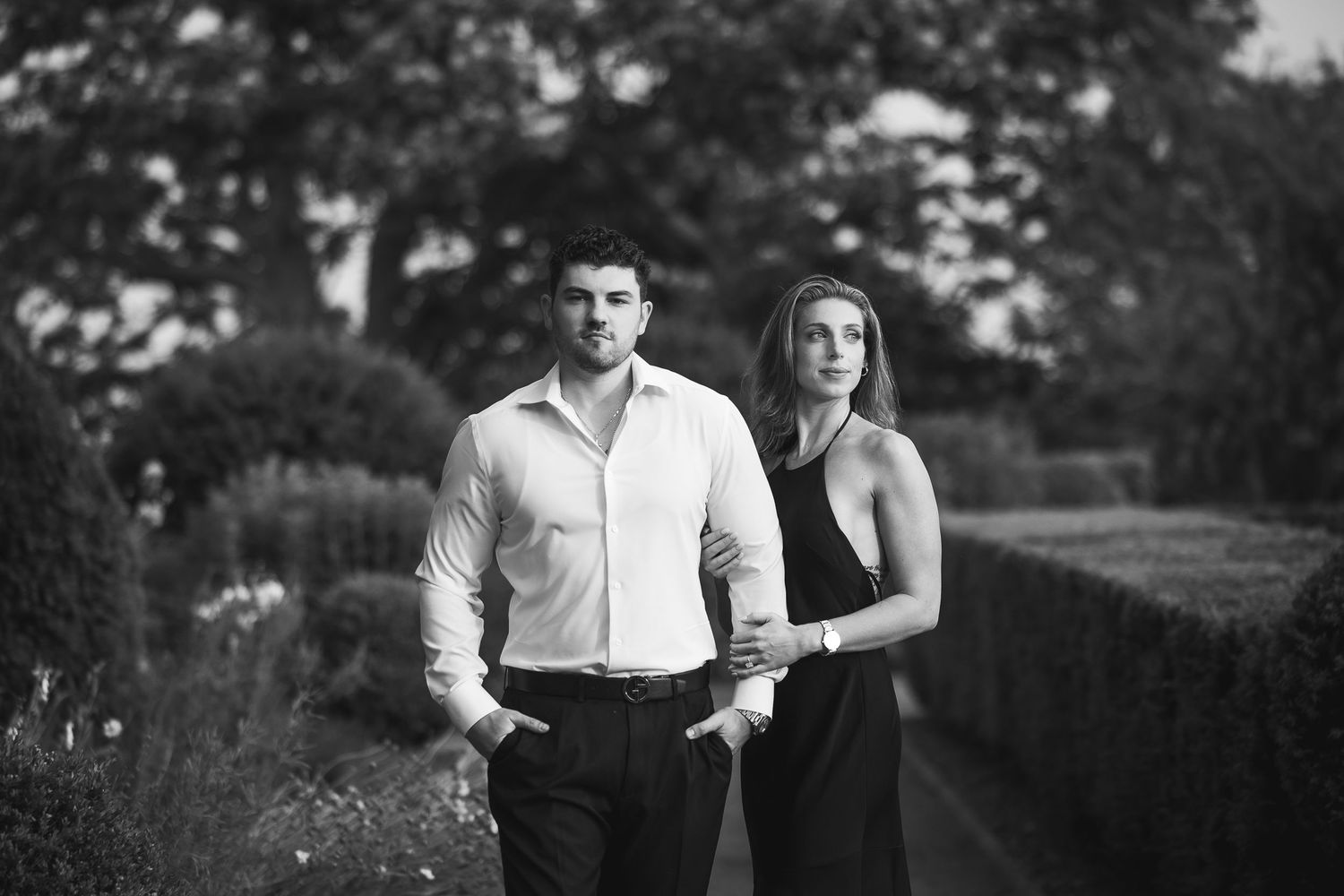 A couple stands close together in a romantic outdoor setting with trees and natural light in black and white.