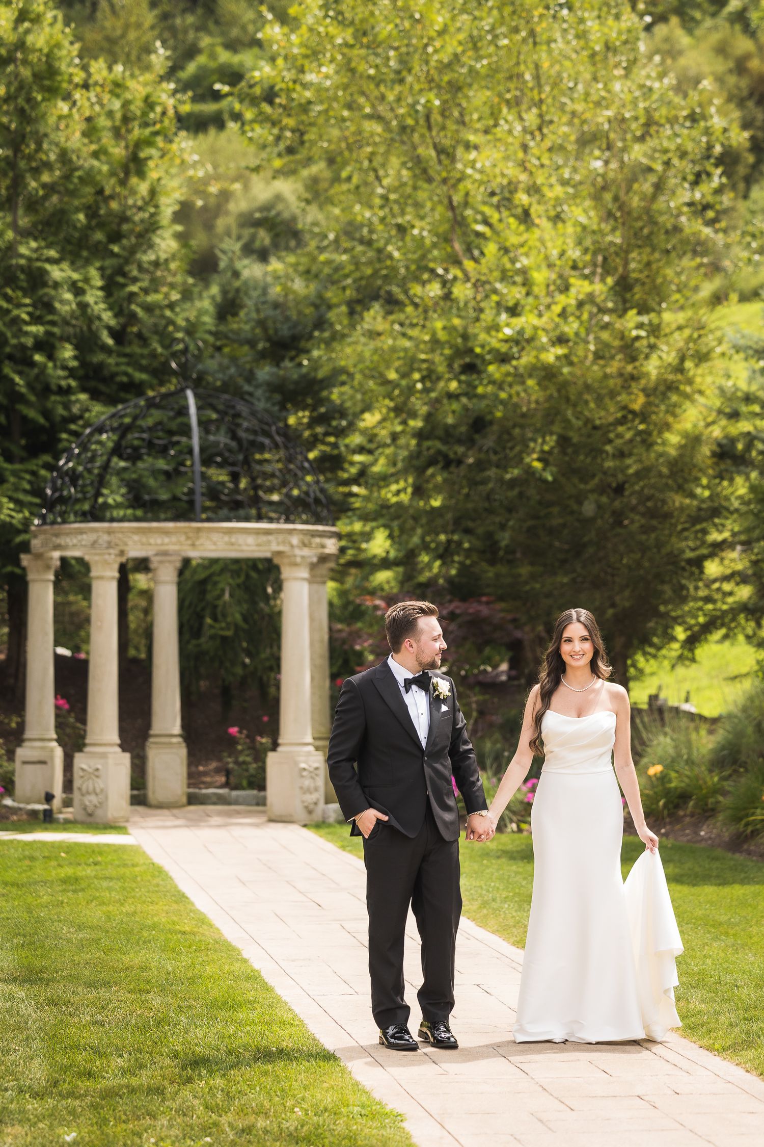 Bride and groom stroll together through elegant outdoor colonnade in garden setting.