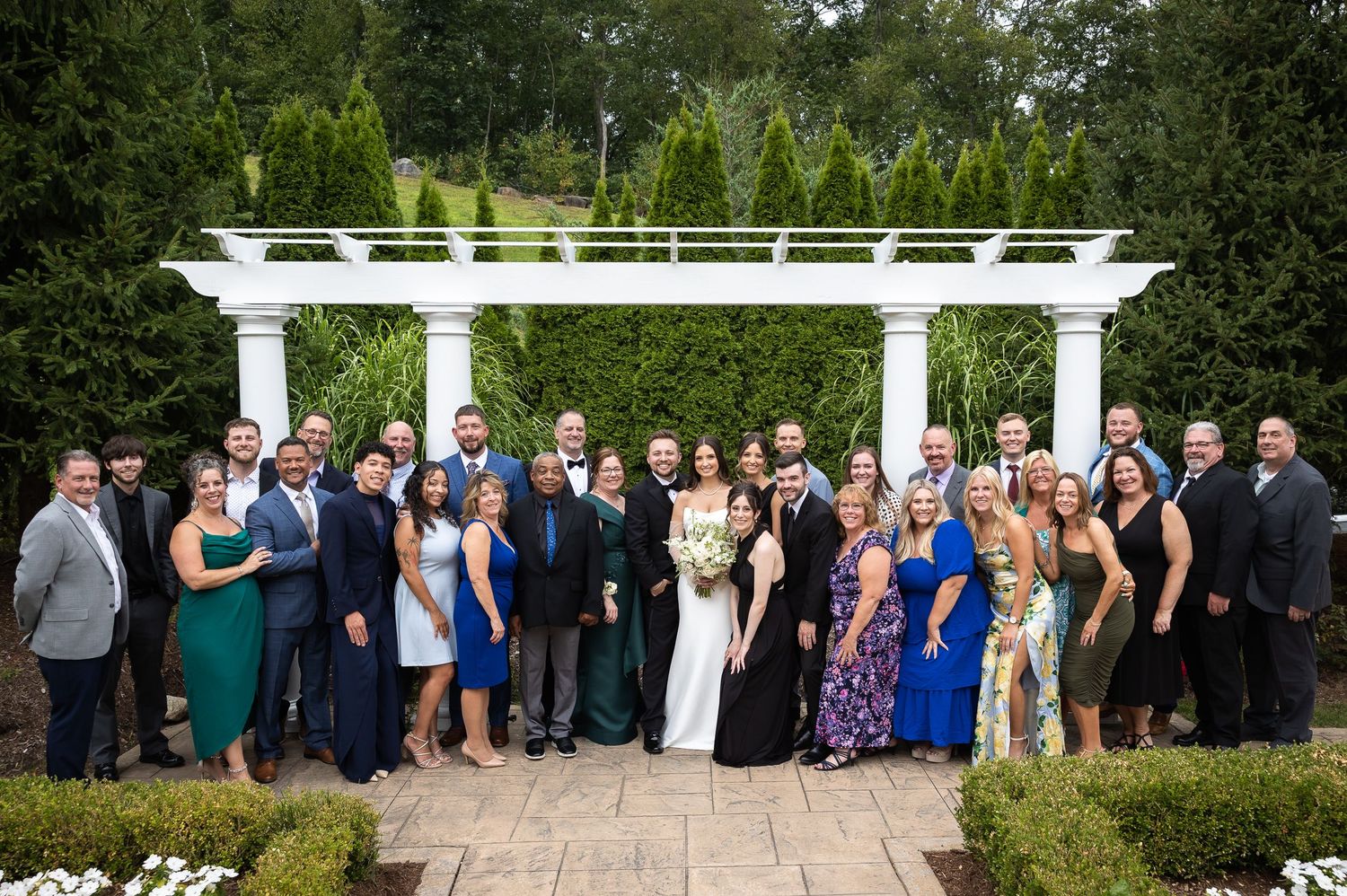 Large wedding party gathered outdoors under a white pergola on a sunny day.