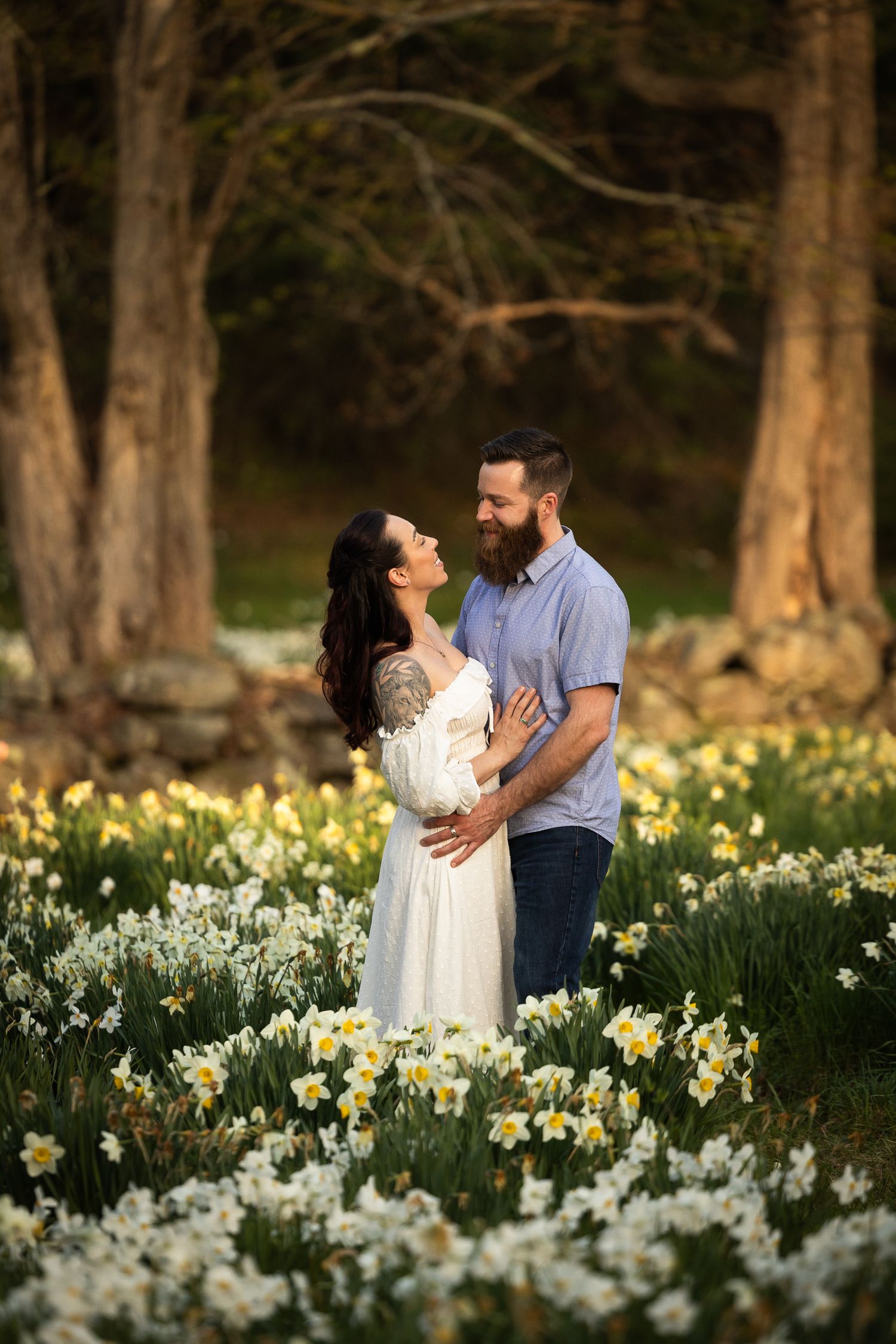 A couple embraces in a scenic garden filled with blooming white daffodils and tall trees in the background.