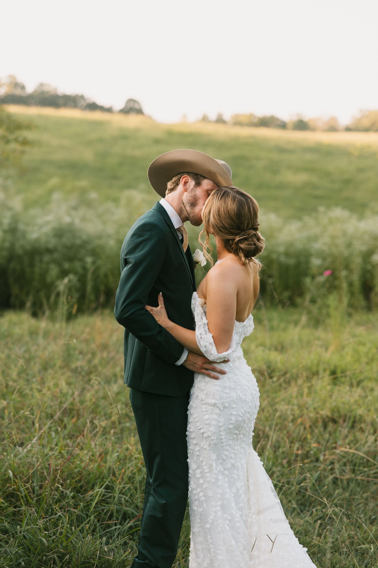 Bride and groom kissing in golden hour meadow, groom in dark suit and cowboy hat, bride in lace gown.