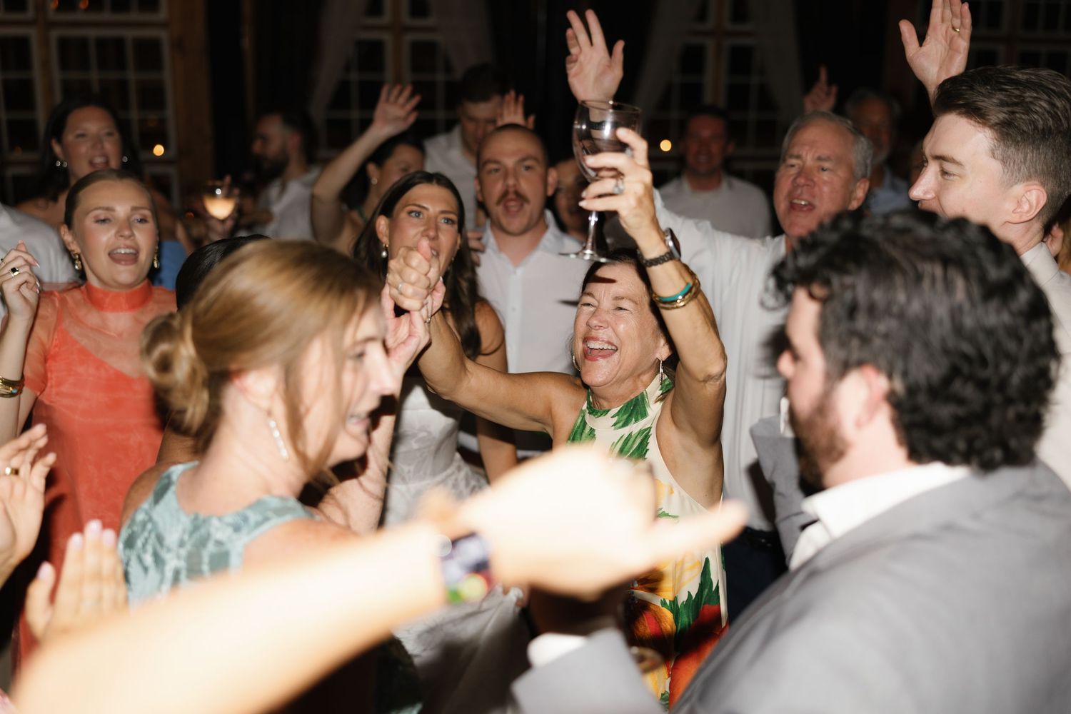 Joyful wedding reception guests dancing and celebrating with raised hands and drinks on the dance floor at night.