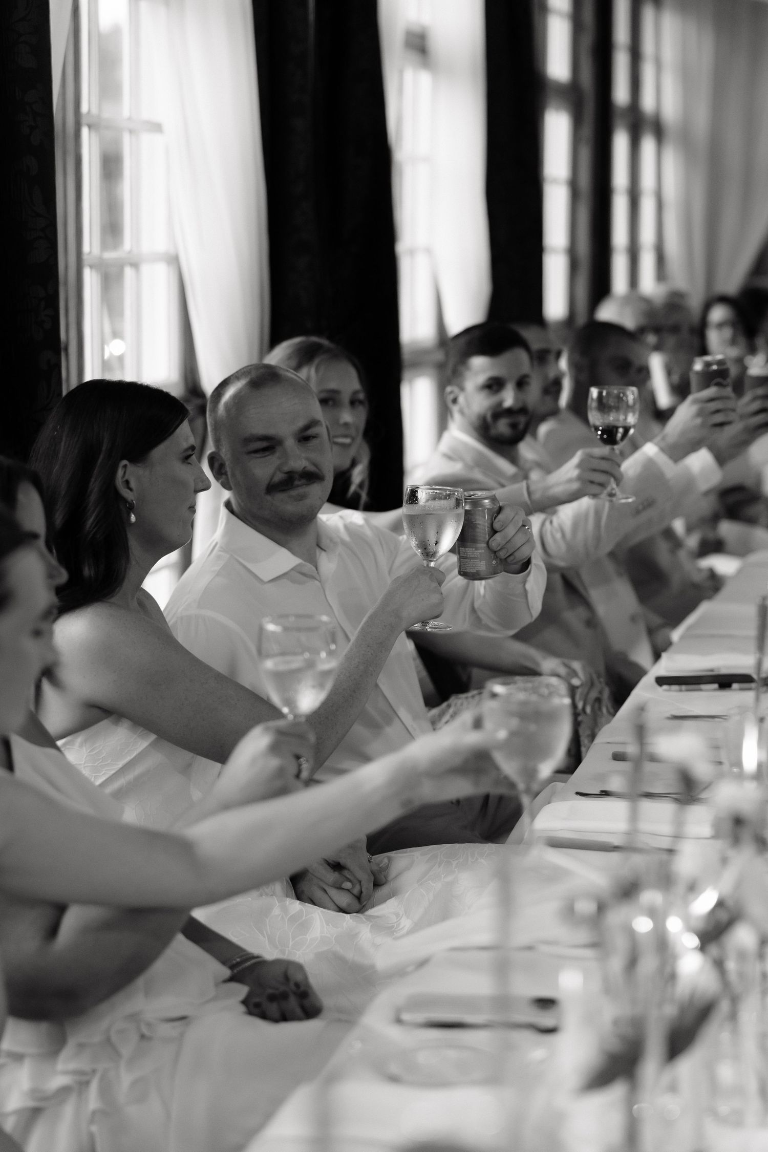 Black and white photo of wedding guests raising glasses in a toast at a elegant reception dinner table.