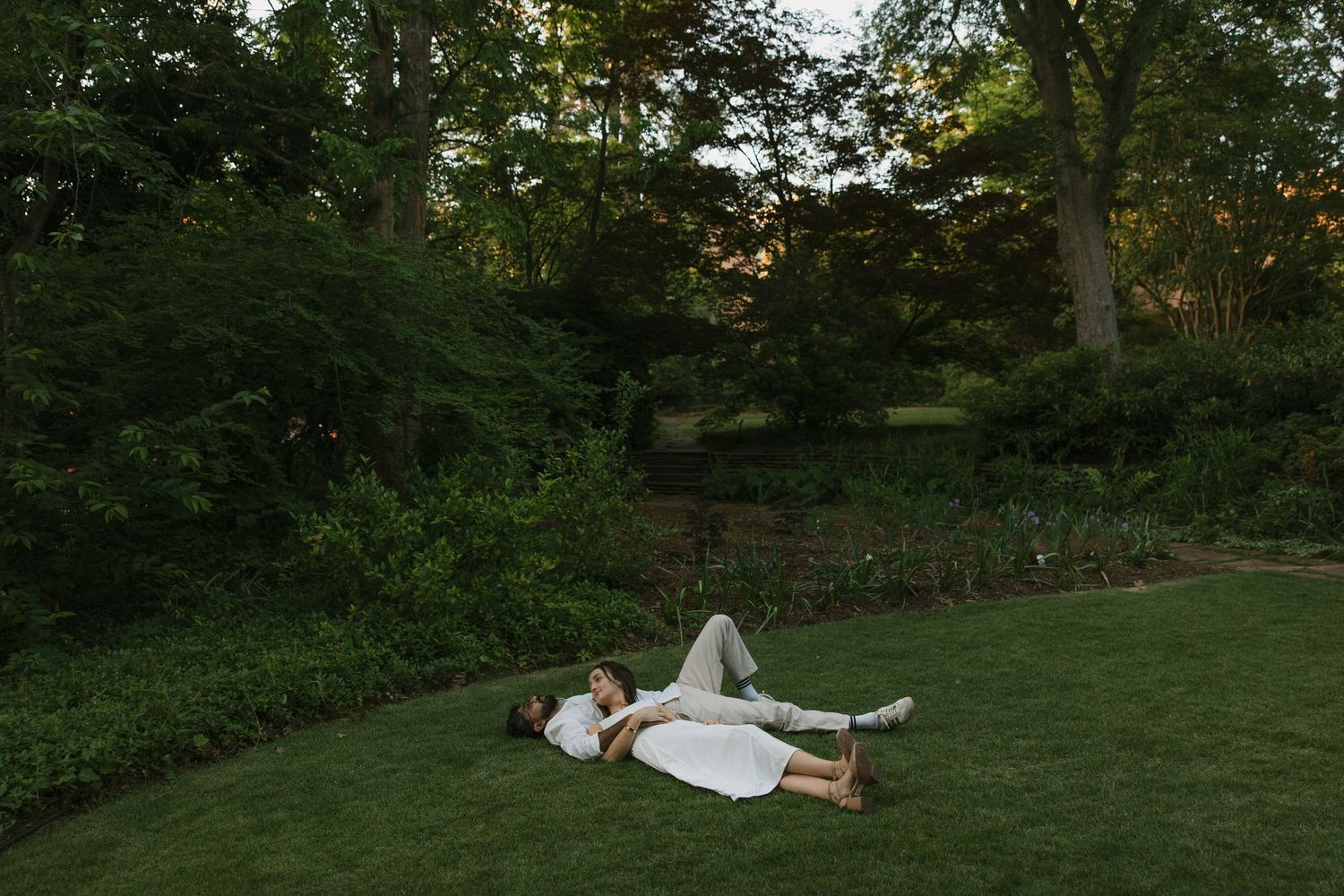 Couple lying on lush green grass in a romantic garden setting surrounded by trees at dusk.