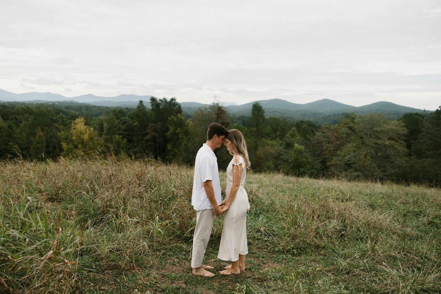 Couple holding hands in a meadow with mountain views and lush green forest in the background on a cloudy day.