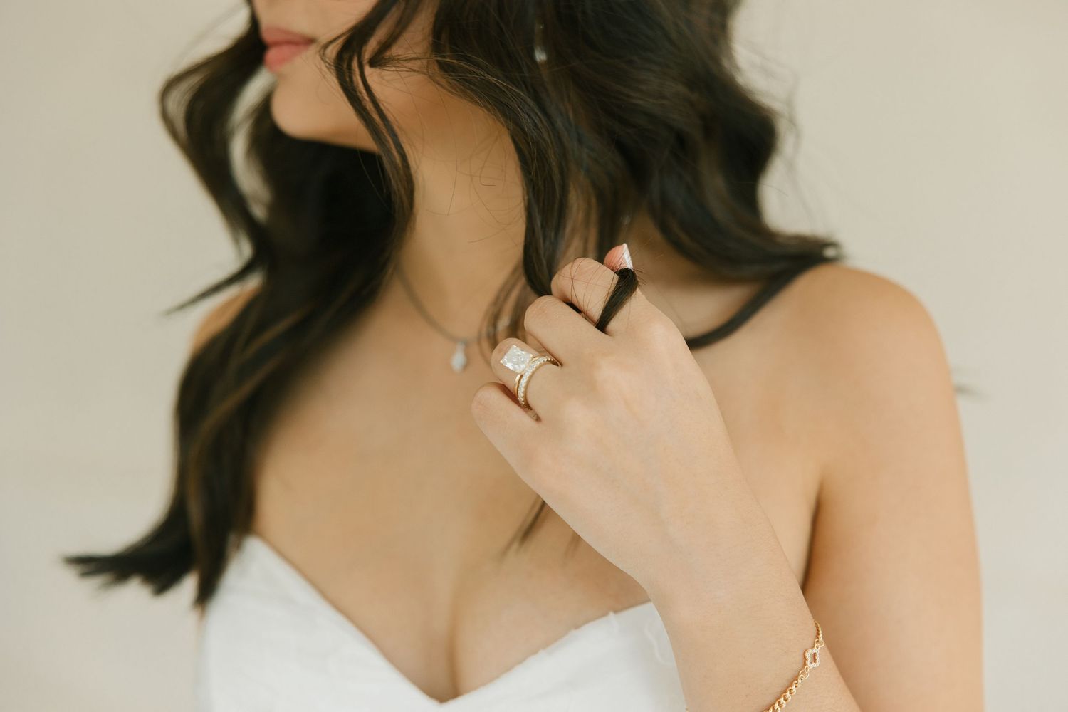 Close-up of bride in white dress touching pearl necklace, wearing stacked rings and gold bracelet.