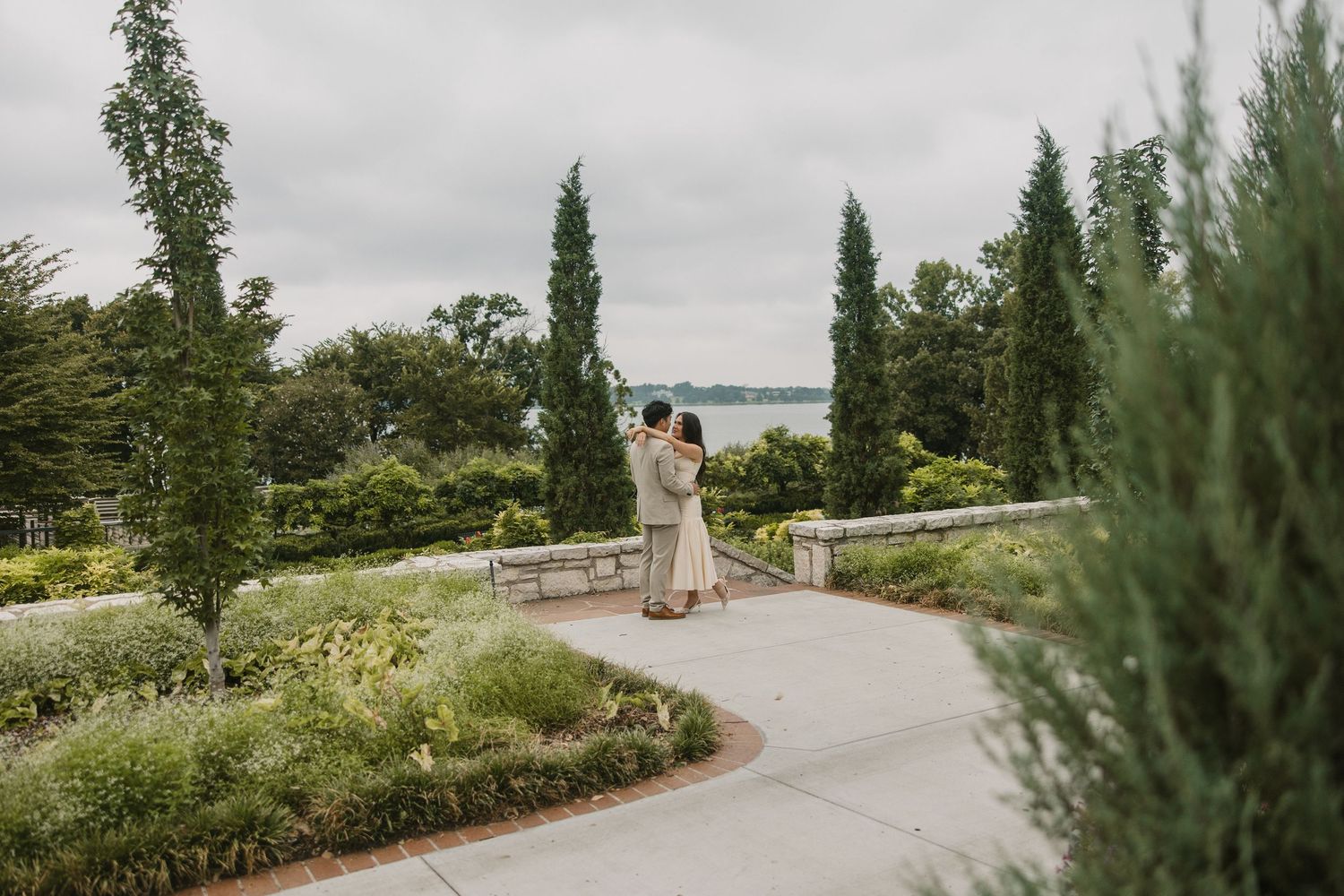 Couple shares tender embrace on curved garden path surrounded by cypress trees and lush greenery.