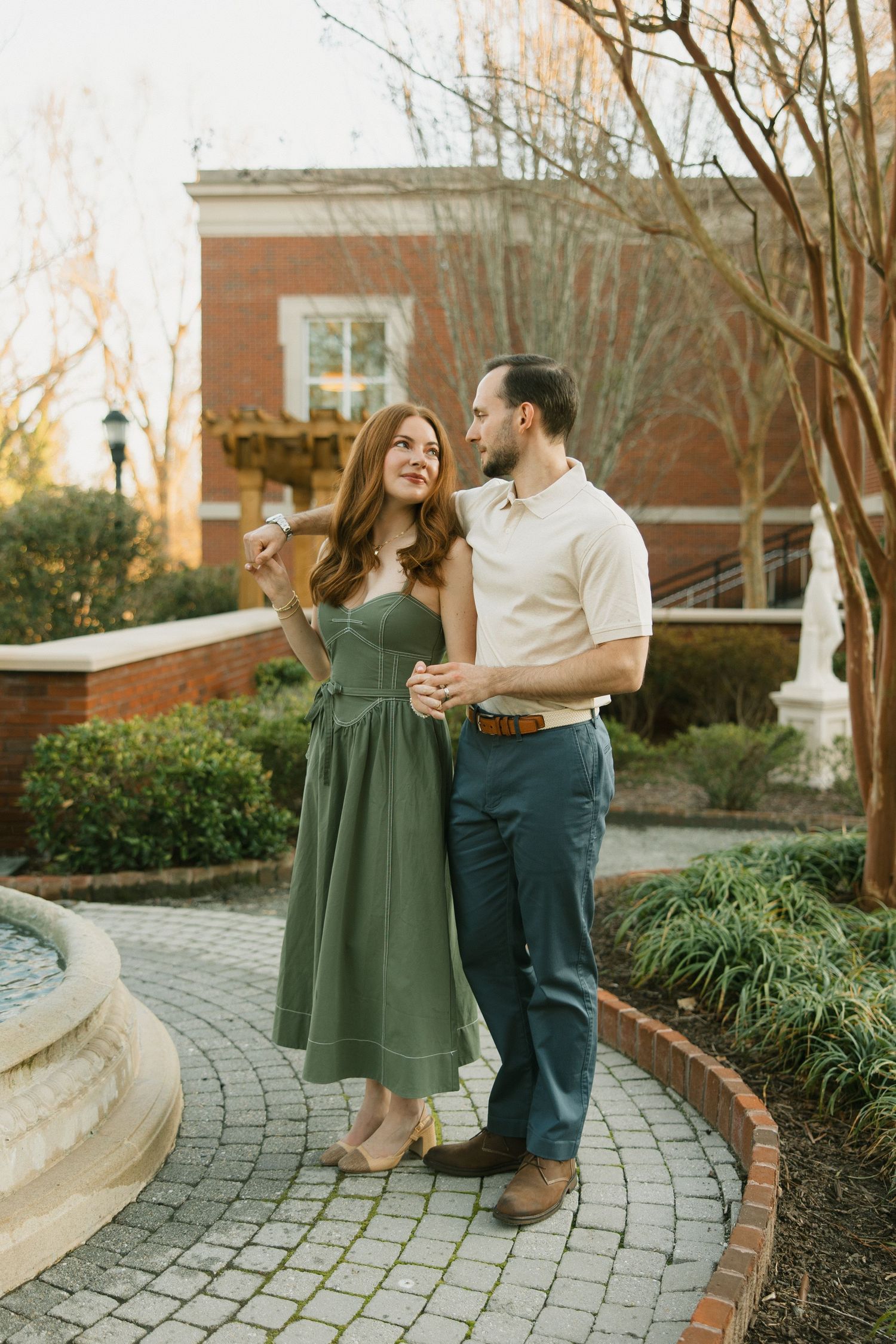 Couple standing together on brick path by fountain, woman in green dress pointing while man in polo smiles at her.