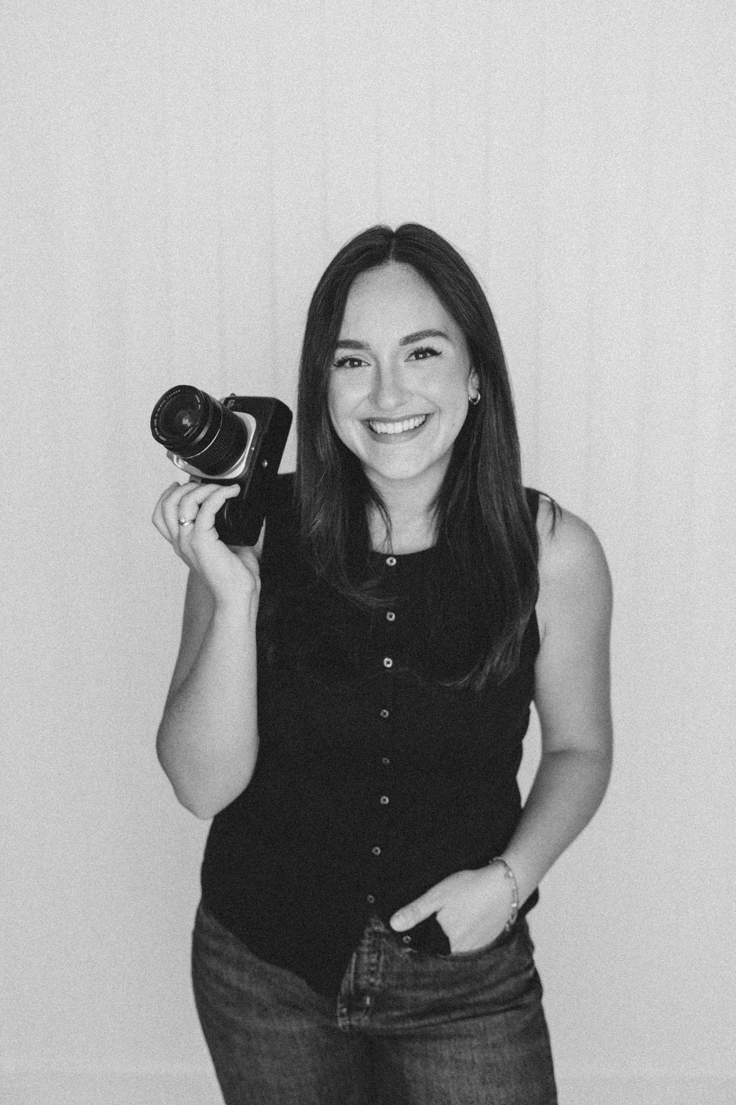 Black and white photo of a smiling female photographer holding a camera in a studio setting.