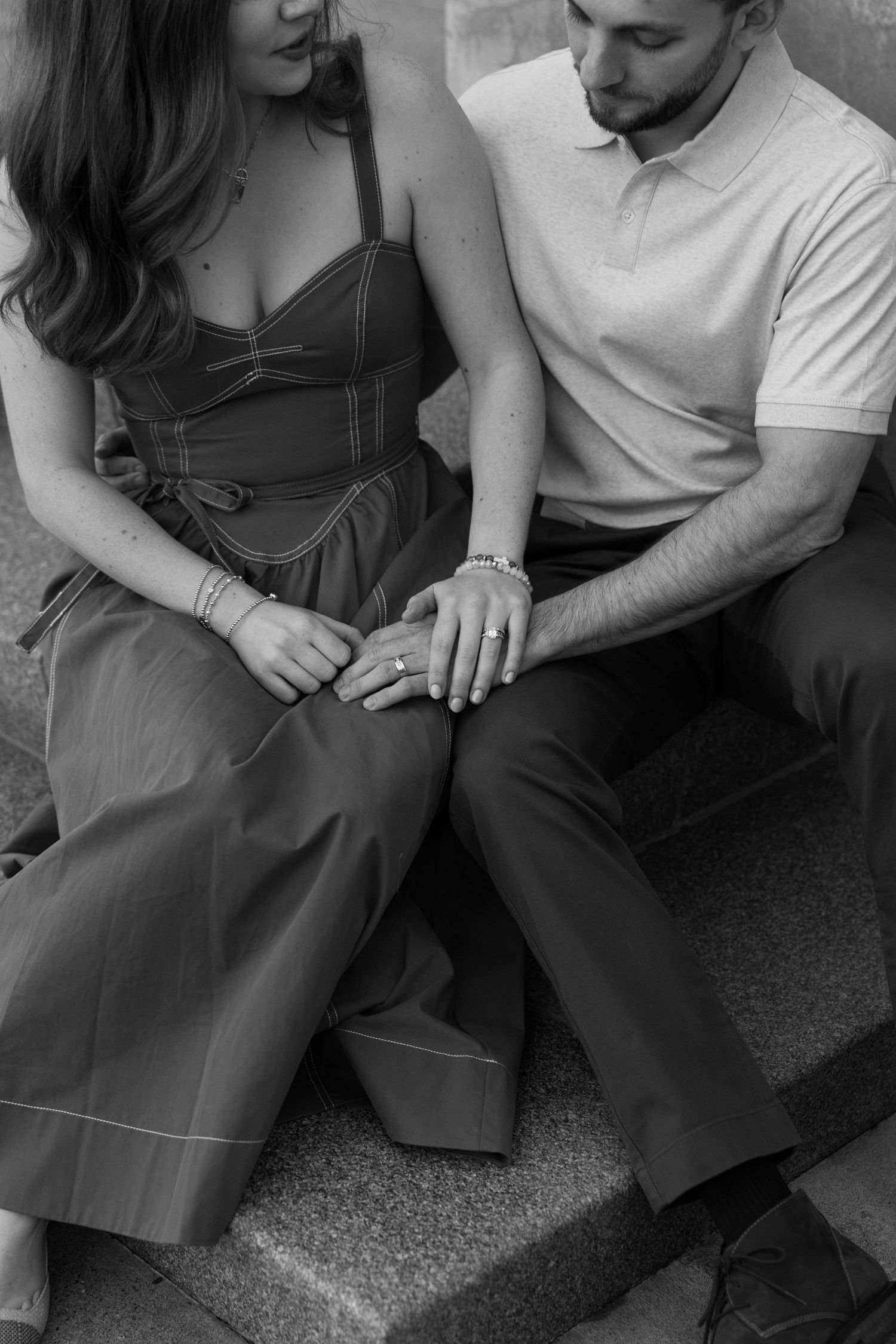 Black and white photo of a couple sitting on steps, holding hands and admiring an engagement ring.