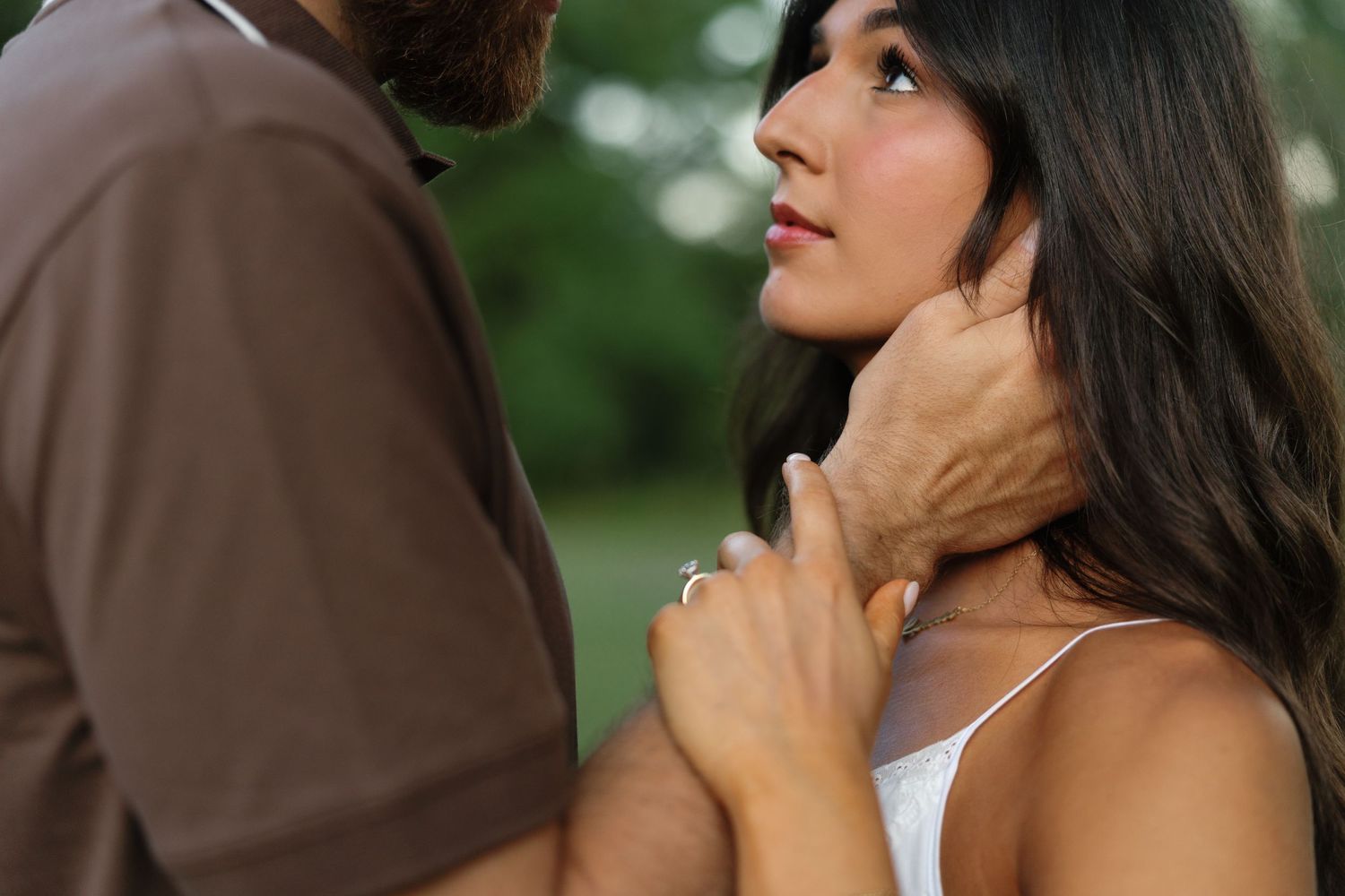 A man tenderly holds a woman's face as she gazes up at him outdoors in a lush green park setting.