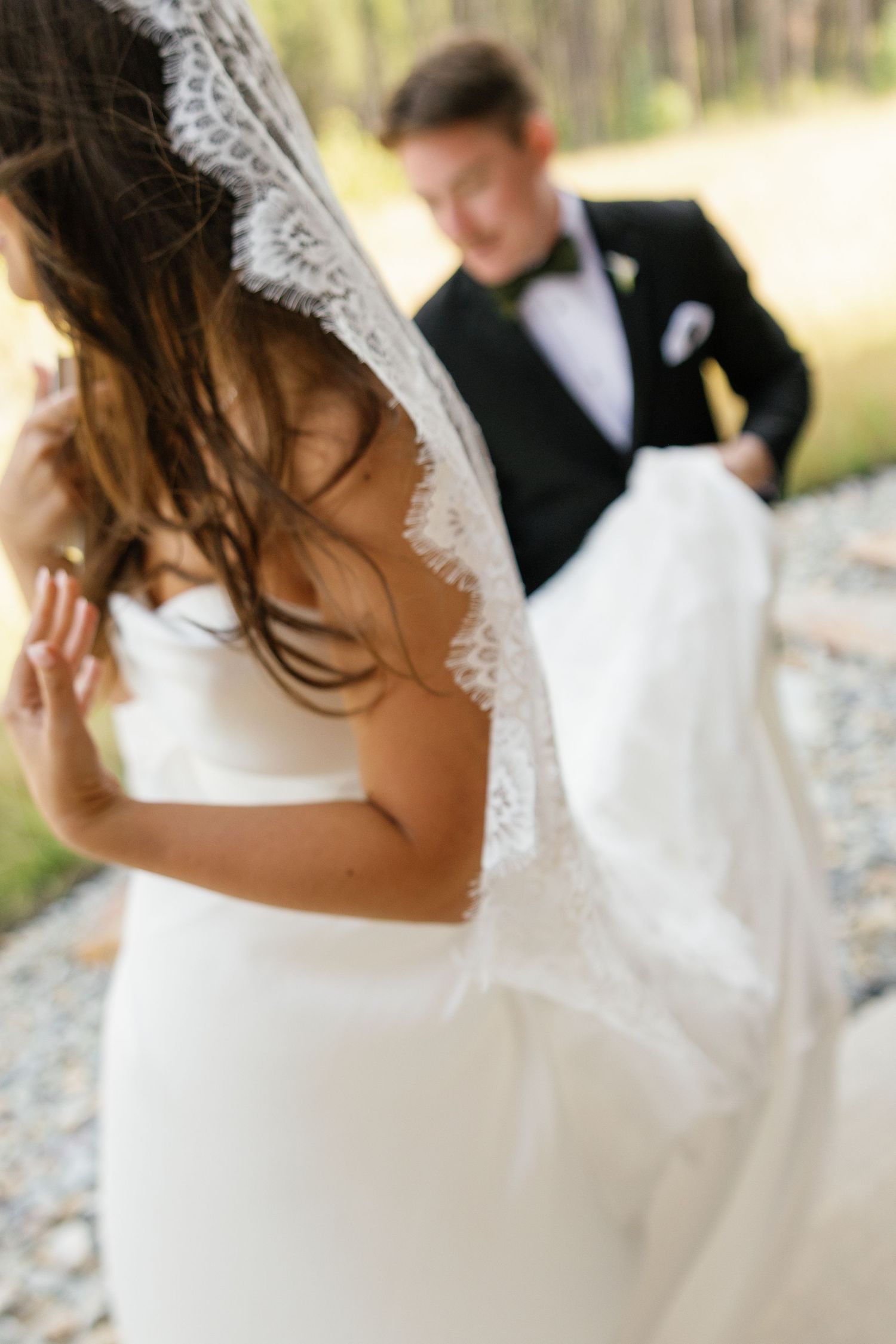 Bride in white strapless gown with lace veil, groom in black tuxedo holding her train outdoors.