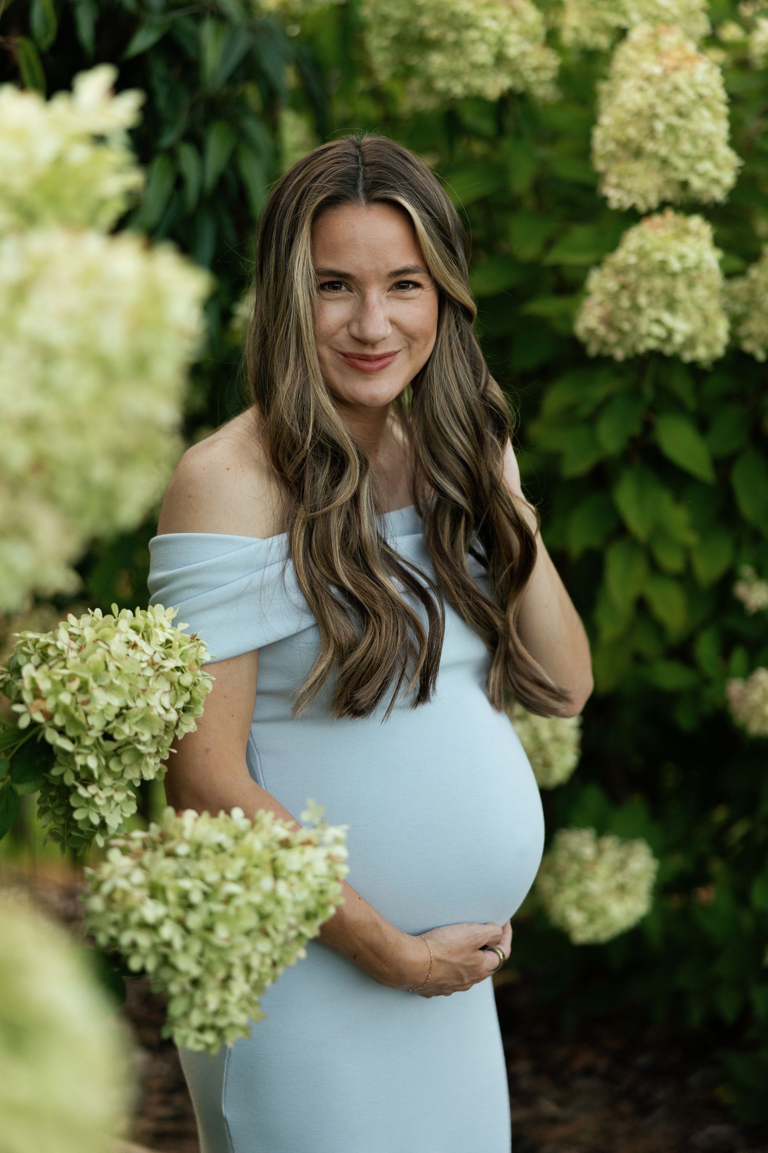 Pregnant woman in light blue off-shoulder dress cradling baby bump among white hydrangeas in lush garden.