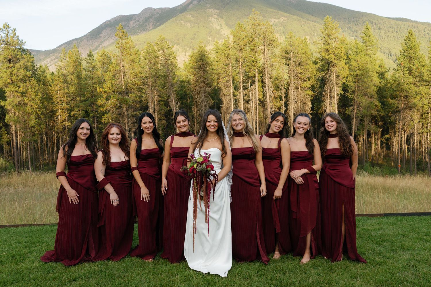 Bride in white gown poses with eight bridesmaids in burgundy dresses before a mountain and forest backdrop.