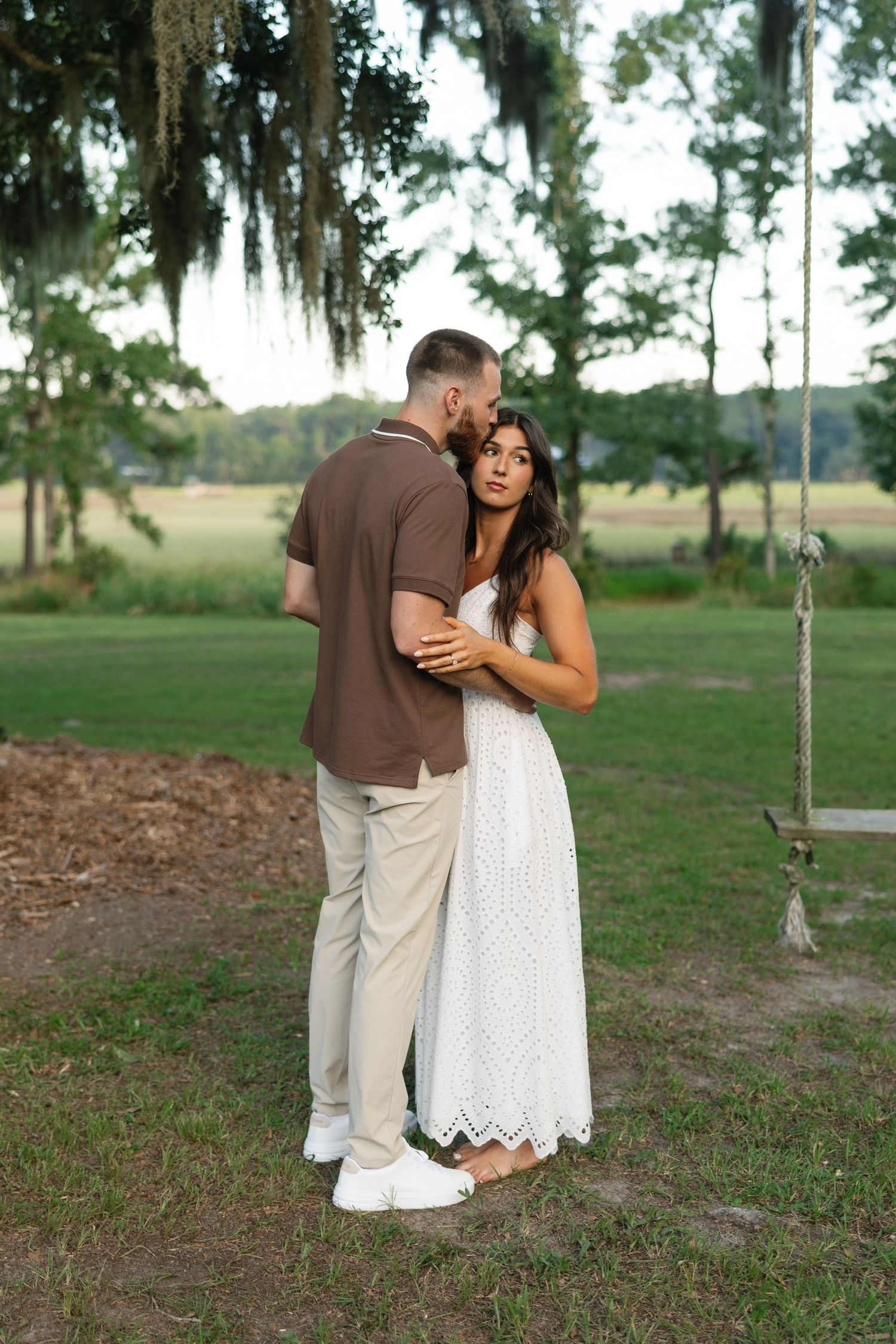 Couple embracing outdoors near a swing, woman in white lace dress, man in brown shirt, surrounded by green trees.