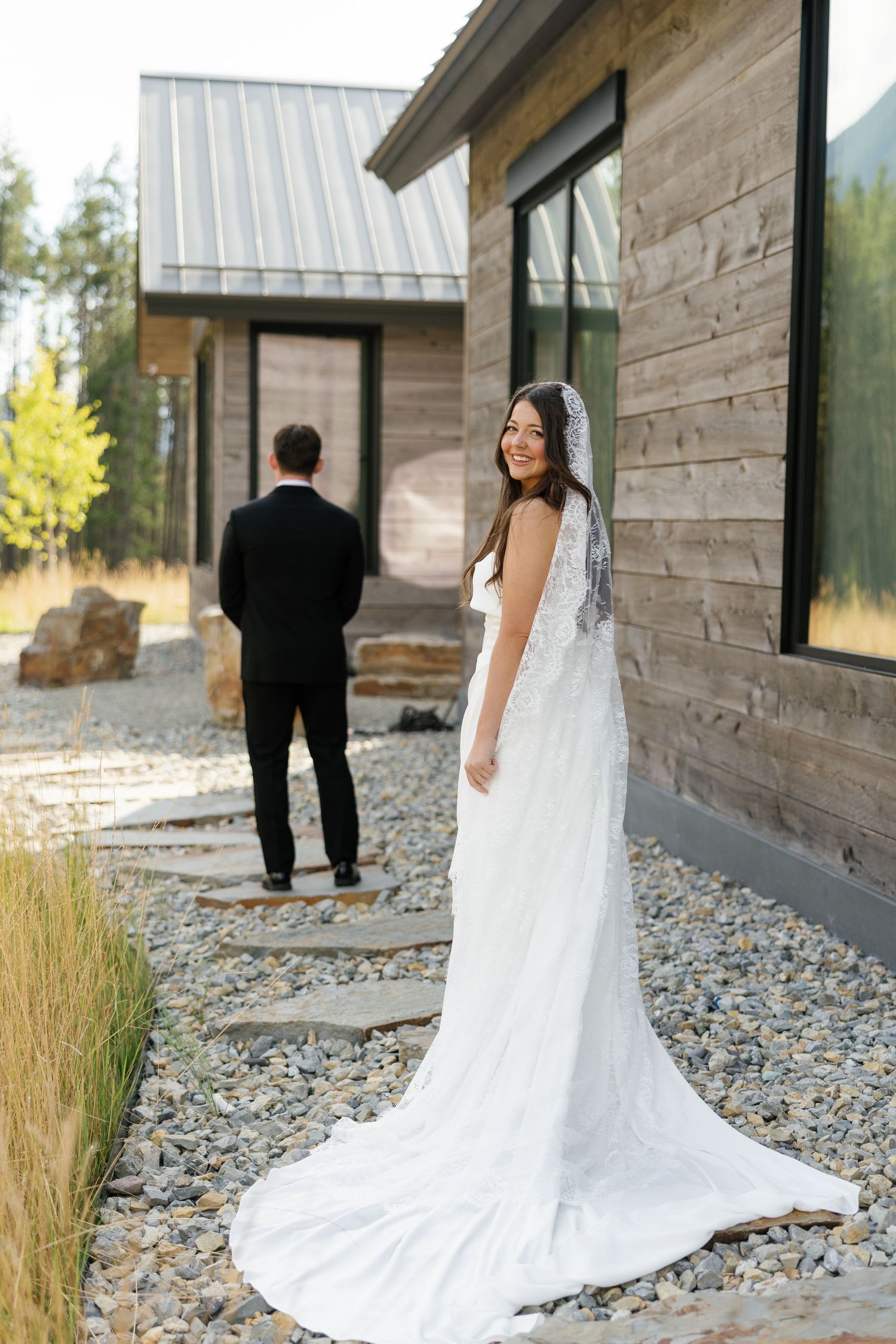 Bride in white lace gown with veil smiles over shoulder as groom stands facing modern wooden building.