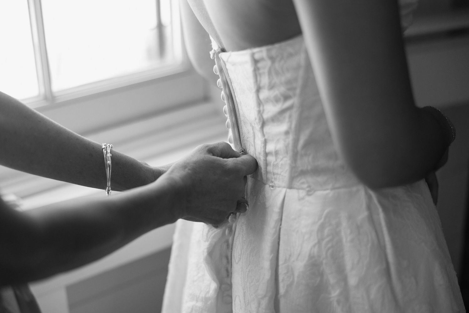 Black and white close-up of hands fastening the back of a white wedding dress by a window.