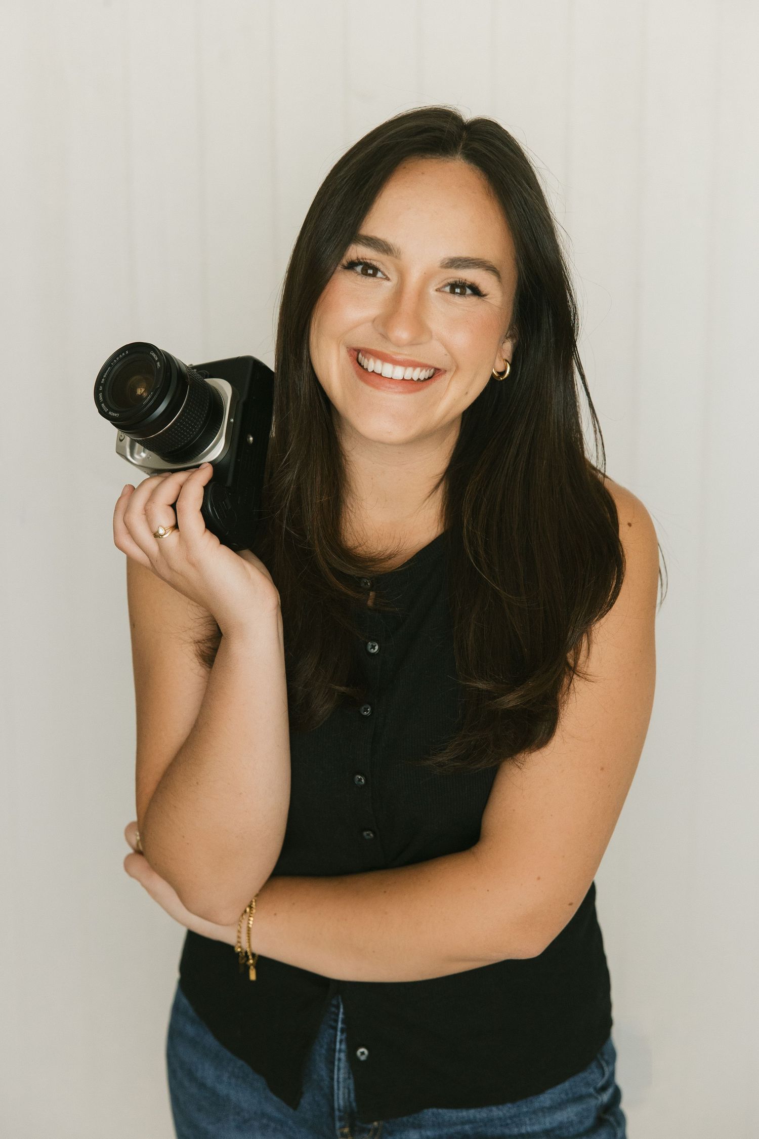 Portrait of a smiling female photographer in a black top holding a camera against a light wall.